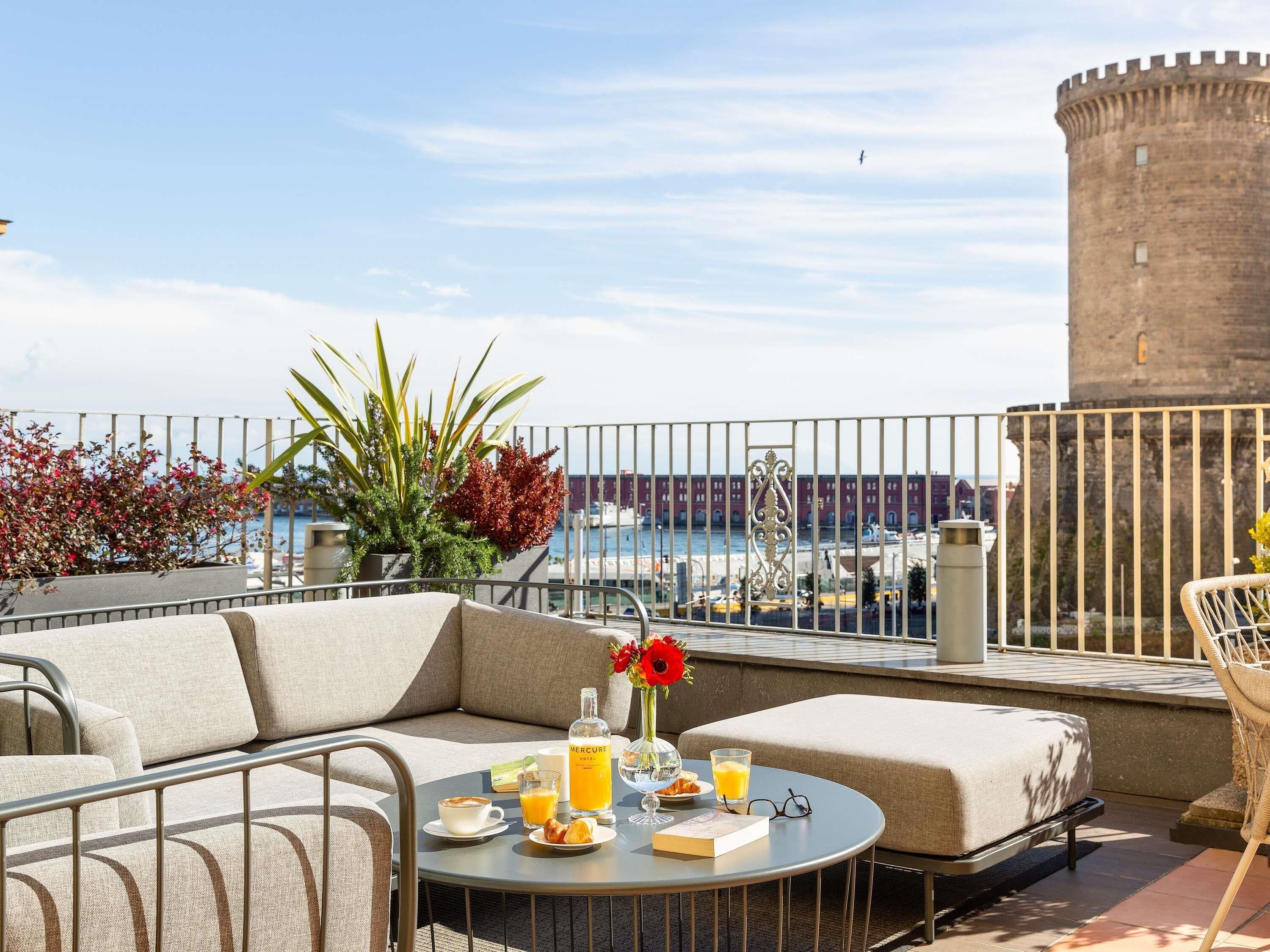 A rooftop terrace with a sofa, a round table with breakfast items, a view of a historic tower, and potted plants.