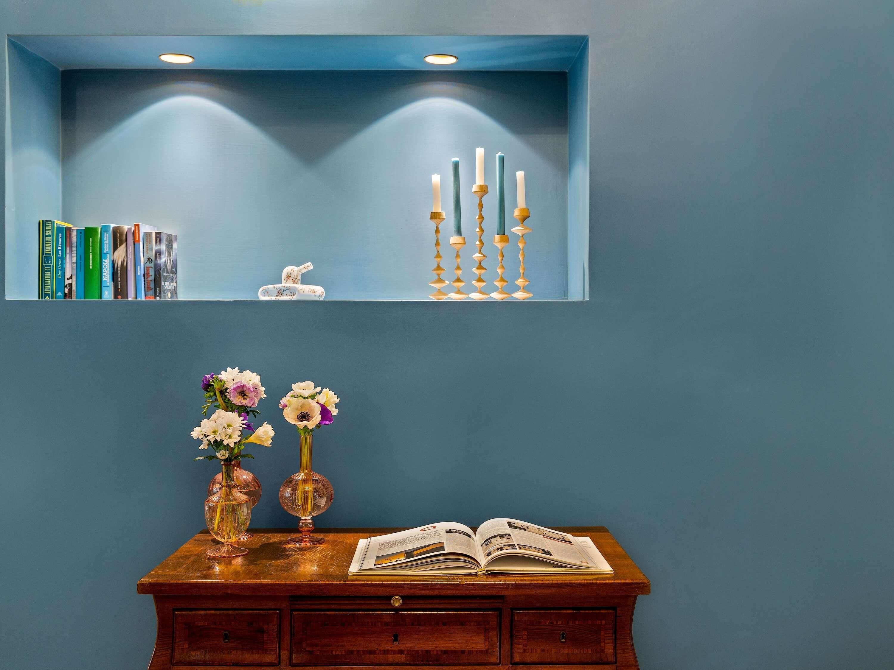 A room with a blue wall, a wooden cabinet with books, a vase with flowers, and a shelf with candles.