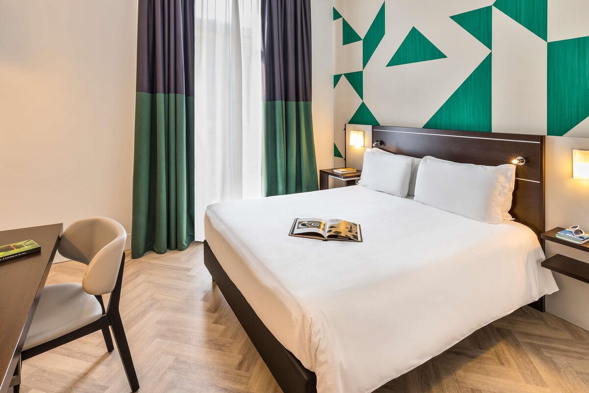 A hotel room with a bed, a chair, a nightstand, and a wall with geometric patterns.