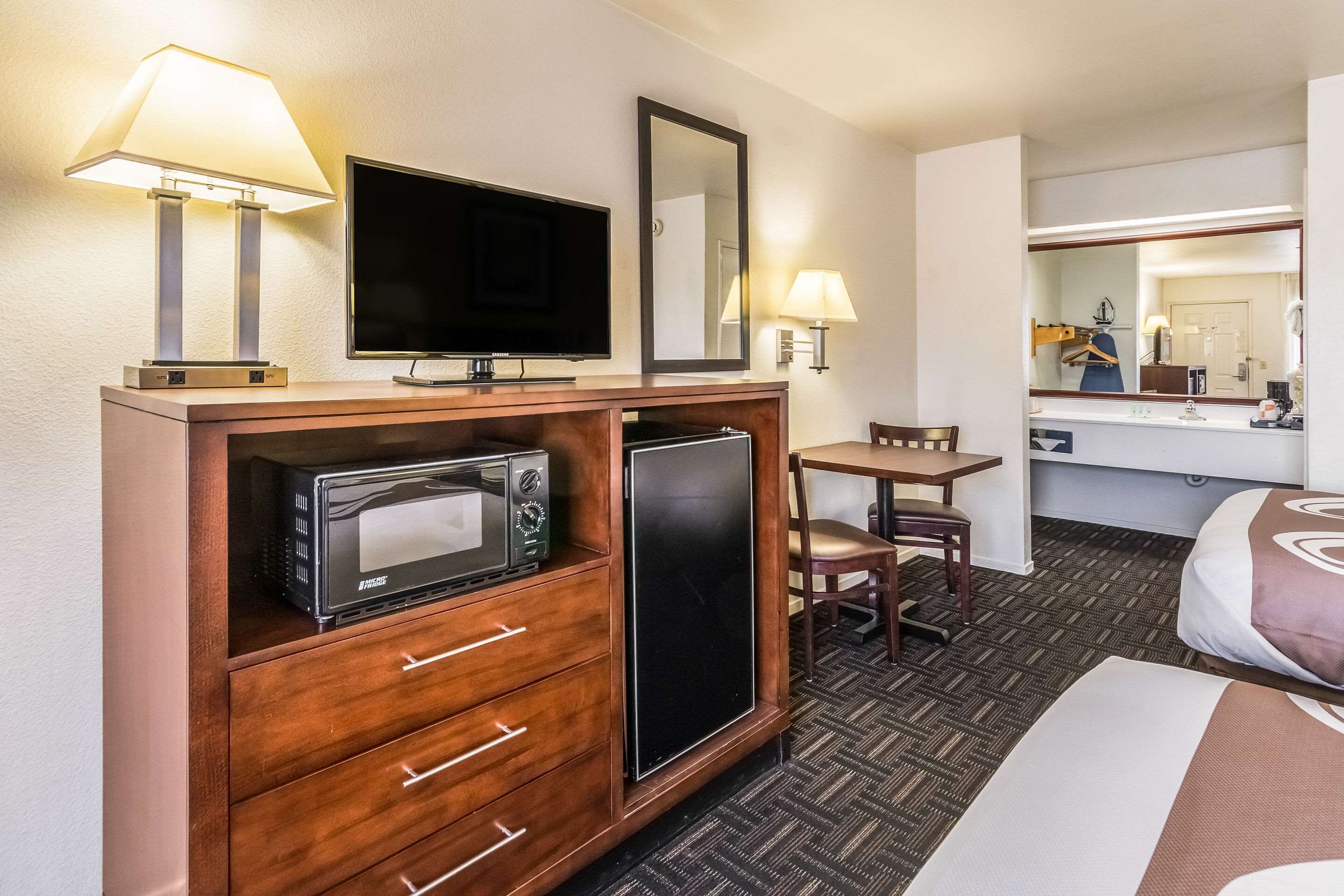 Une chambre d’hôtel équipée d’une télévision, d’un four à micro-ondes, d’un réfrigérateur, d’une commode en bois et d’une petite table avec une chaise.