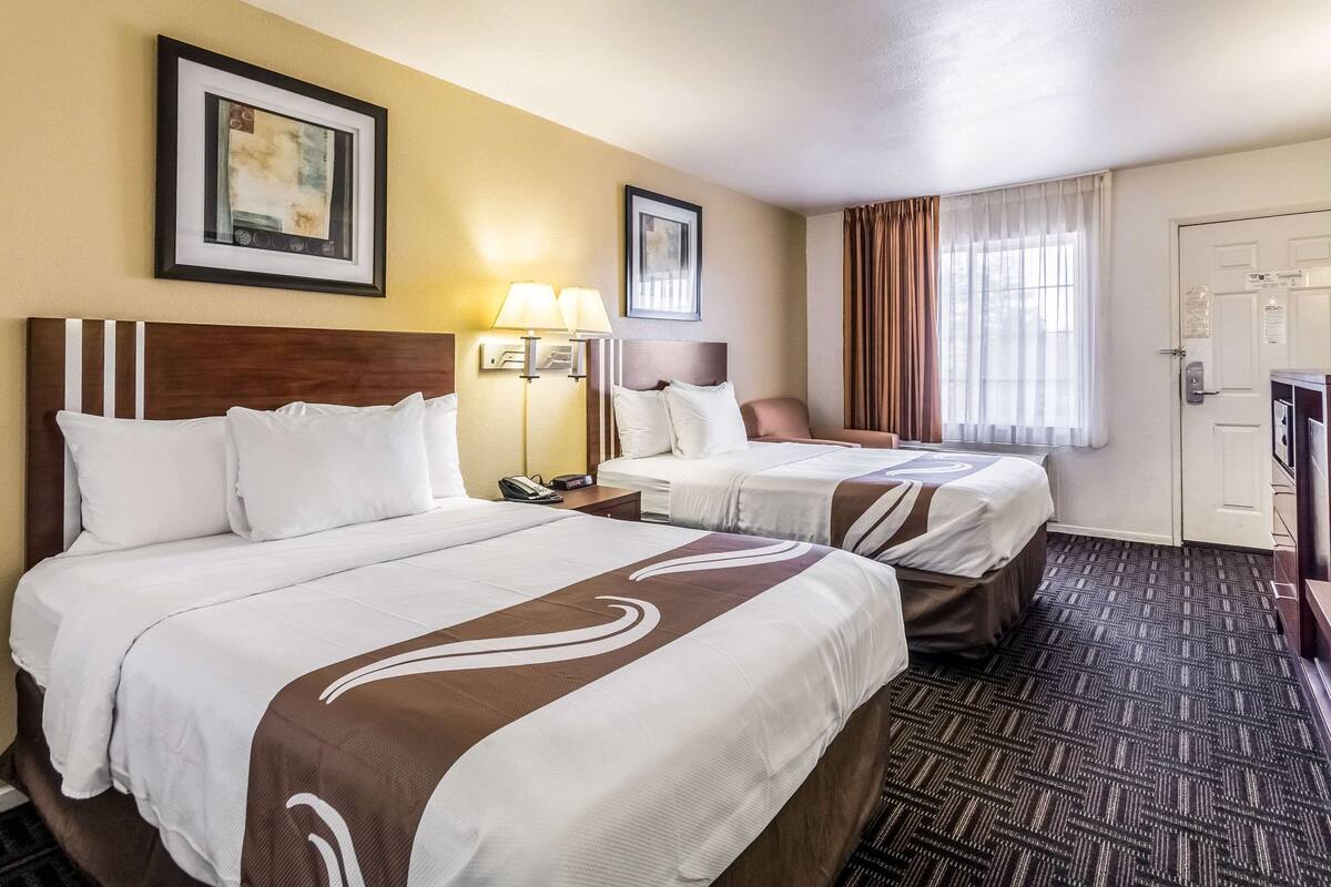 A hotel room with two beds, a desk, a chair, a lamp, and a door.