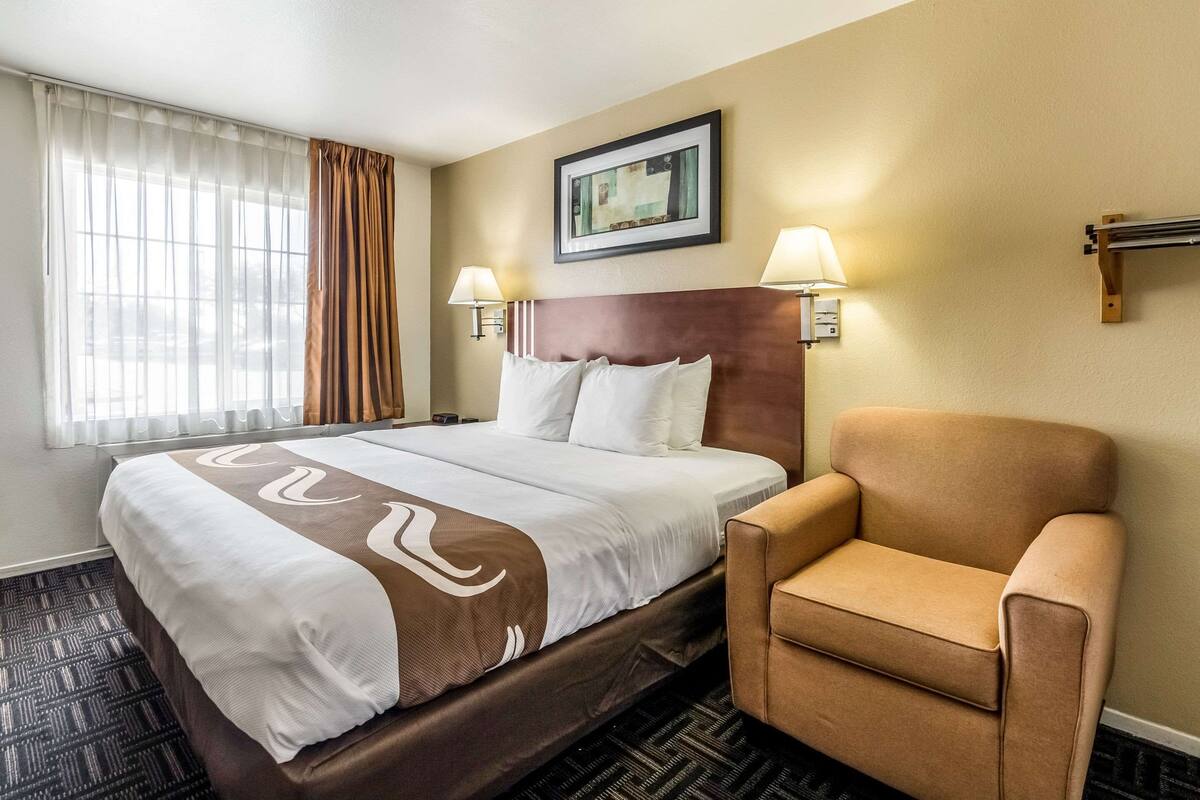 A hotel room with a large bed, two bedside lamps, a brown armchair, a framed picture on the wall, and a window with curtains.