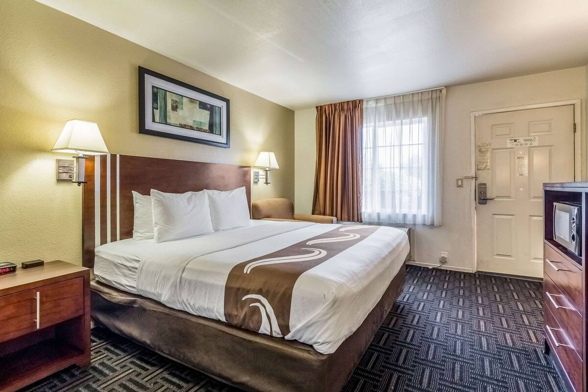 A hotel room with a large bed, two bedside lamps, a nightstand, a dresser, and a door.