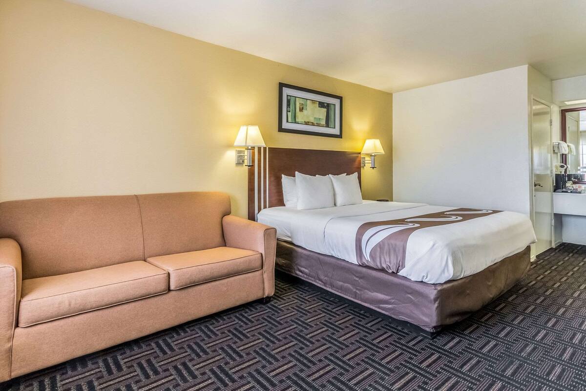 A hotel room with a bed, a sofa, two wall-mounted lamps, a framed picture on the wall, and a carpeted floor.