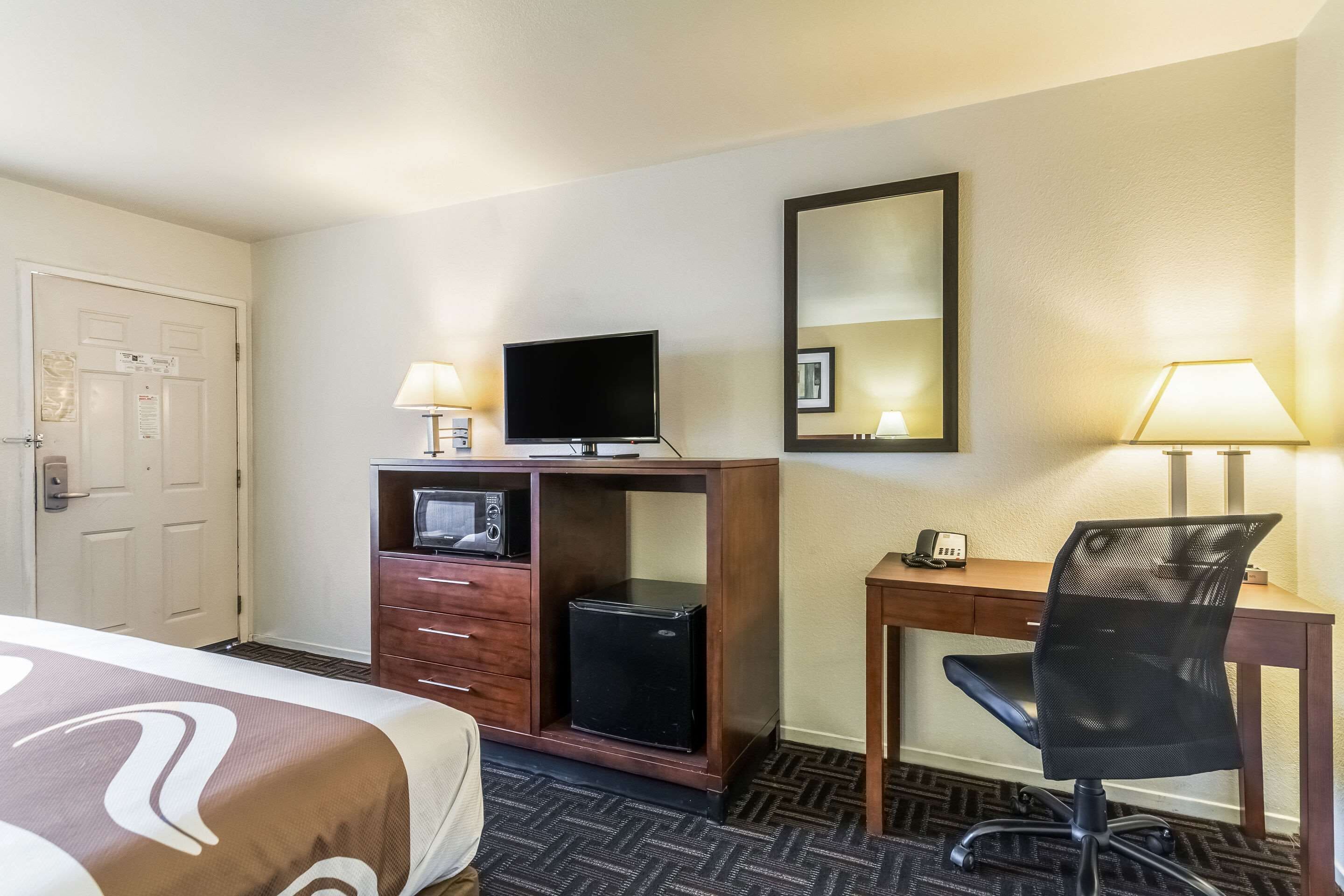A hotel room with a bed, a desk with a computer and a chair, a television, and a small refrigerator.