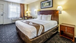 Una camera d'hotel con un letto grande, due lampade sui comodini, una testiera, un quadro incorniciato appeso al muro e pavimento con moquette.