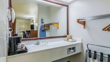 Un baño de hotel con un espejo grande, un lavamanos con artículos de aseo, un perchero y una zona de ducha.