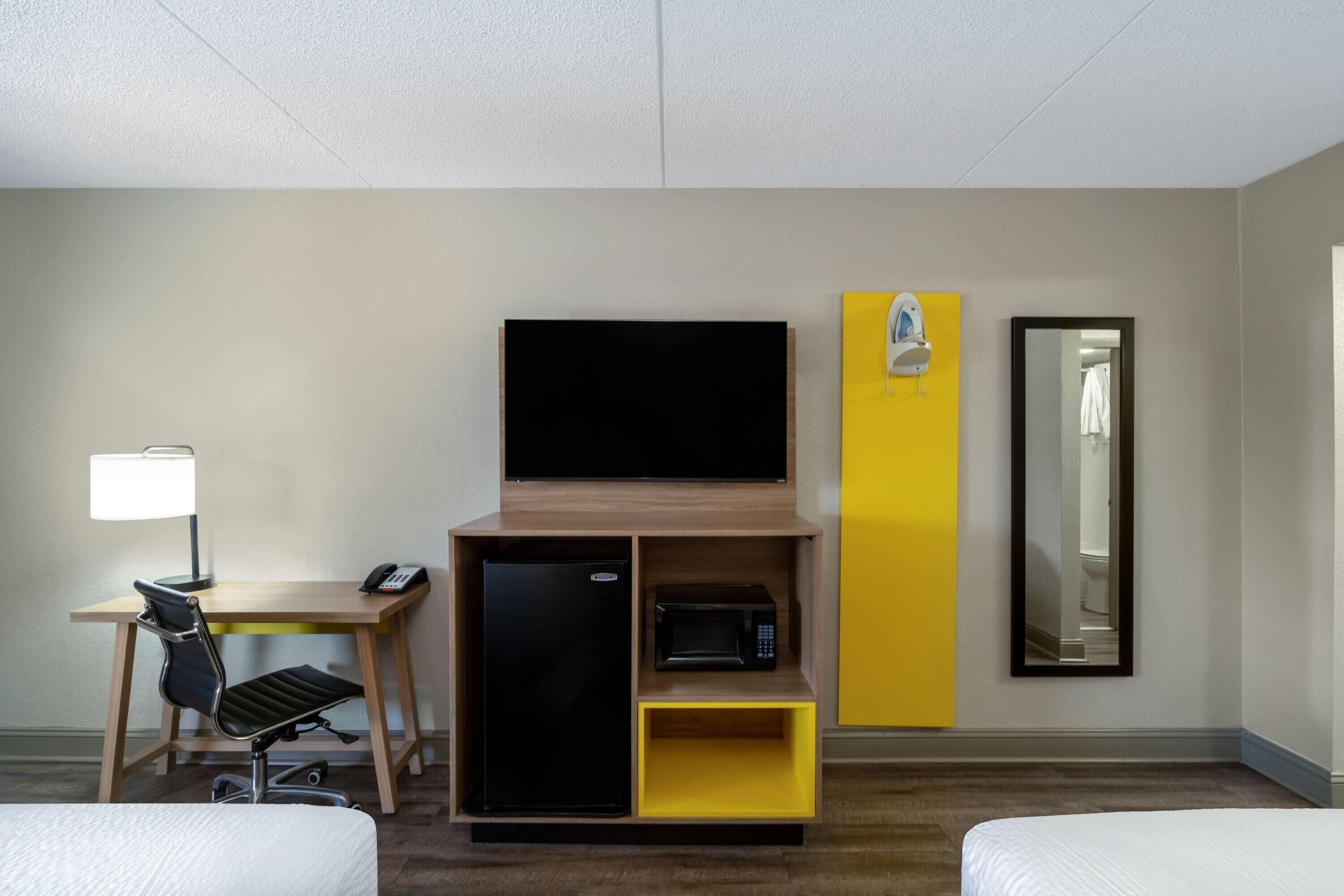 A hotel room with a bed, a desk with a lamp, a refrigerator, a microwave, a television, a mirror, and a yellow cabinet.