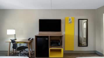 A hotel room with a bed, a desk with a lamp, a refrigerator, a microwave, a television, a mirror, and a yellow cabinet.