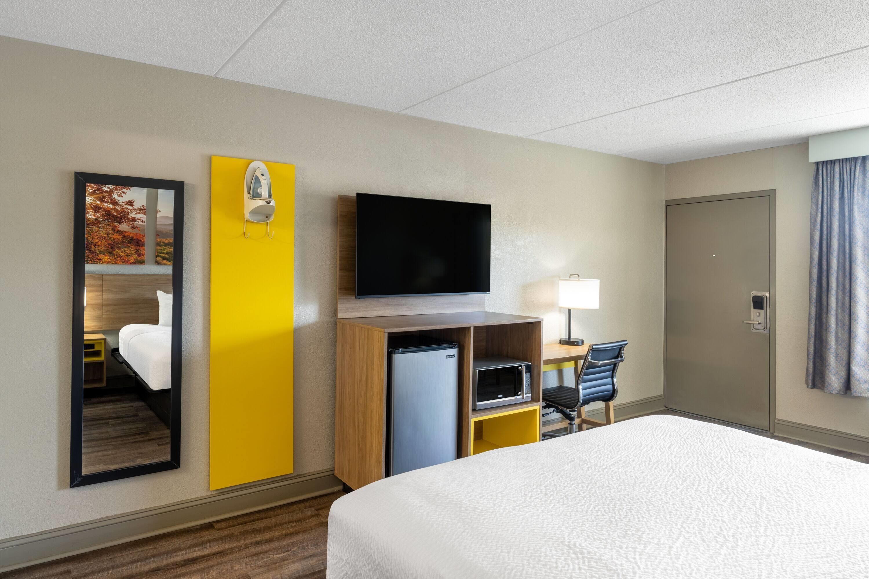 A hotel room with a bed, a flat-screen TV, a microwave, a refrigerator, a desk with a chair, and a mirror.