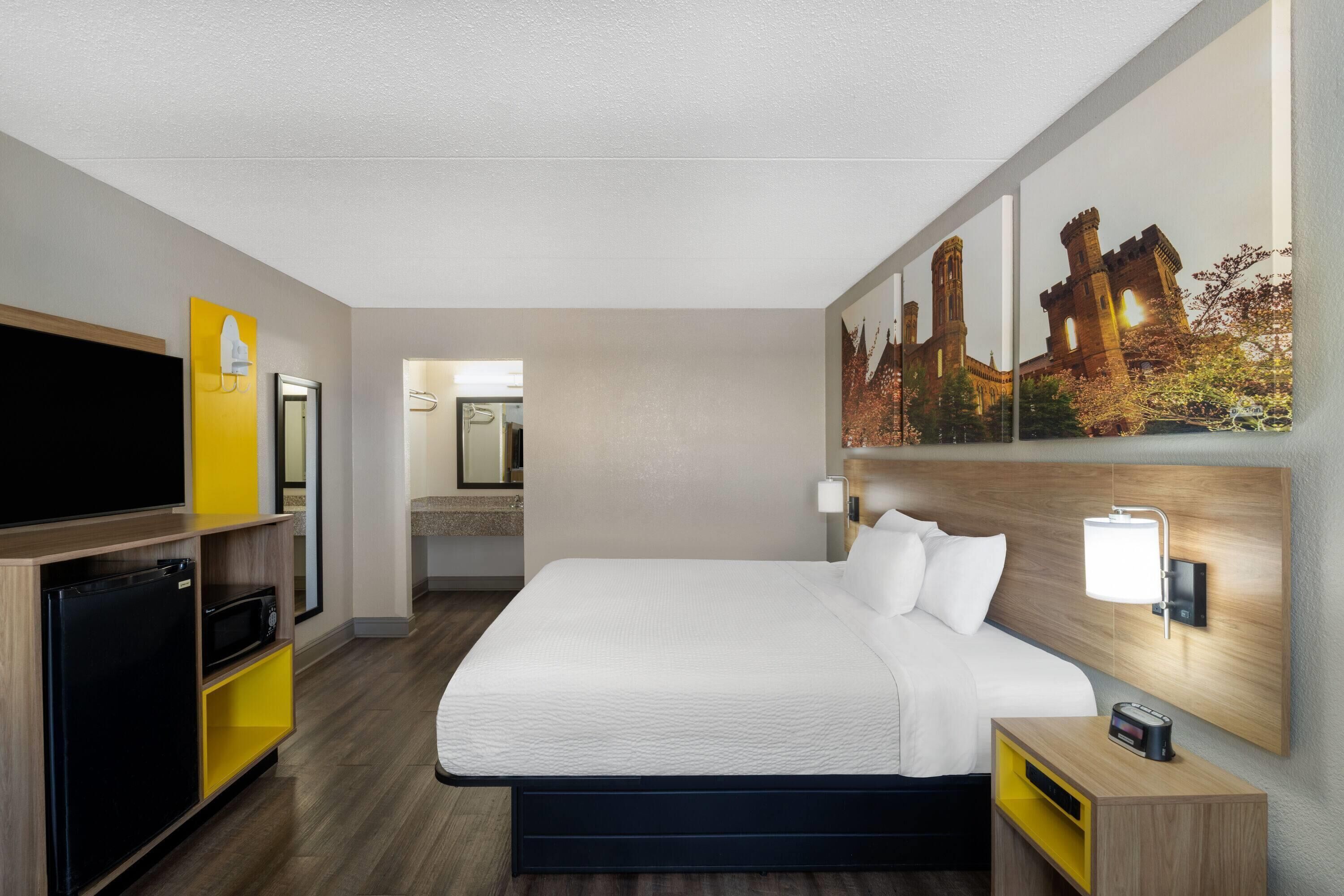 Habitación de hotel con cama, televisión, microondas, refrigerador pequeño, mesita de noche y lámpara de pared.