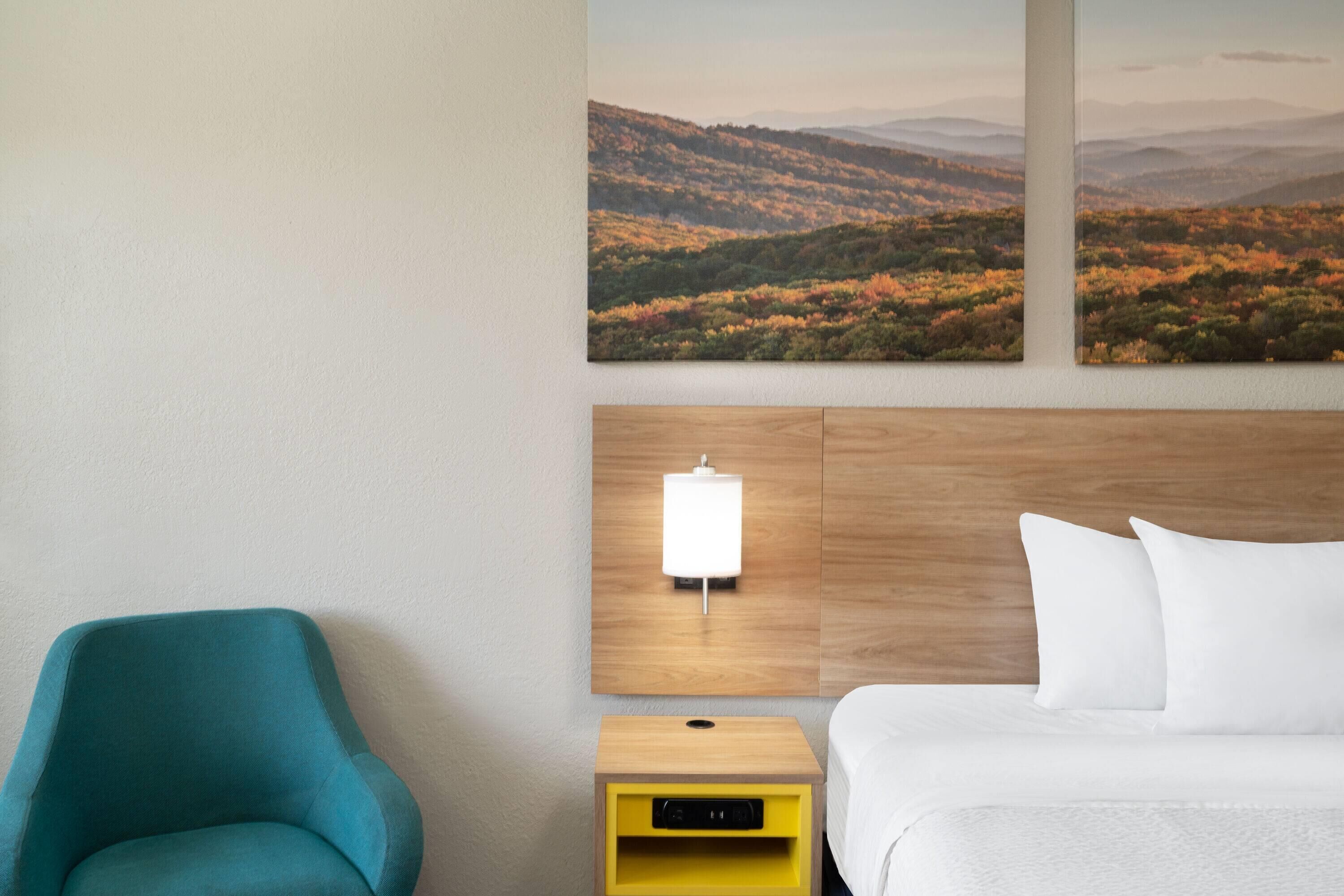 Ein Hotelzimmer mit einem Bett, einem Nachttisch, einer Lampe, einem Sessel und einem Landschaftsbild.