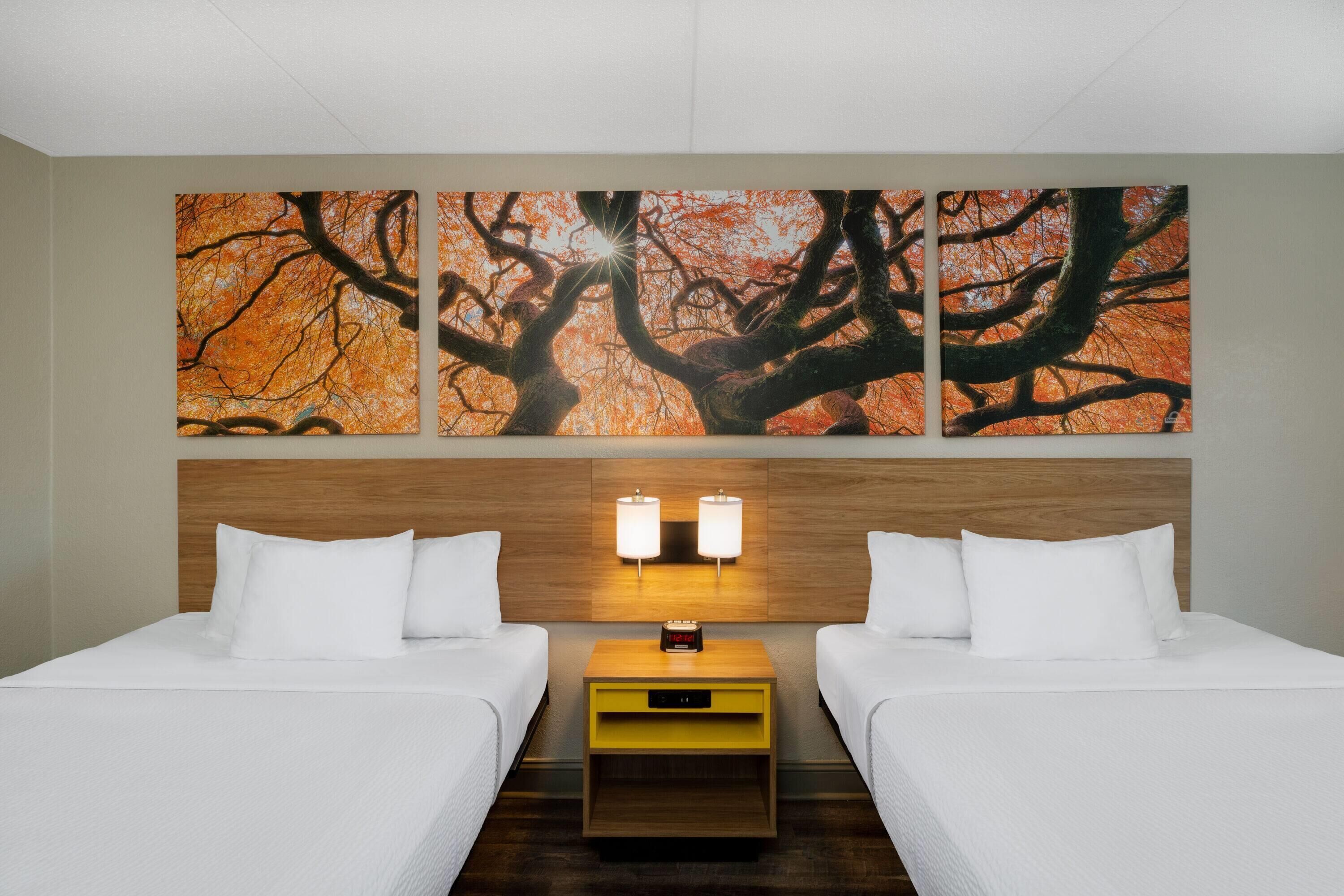 Ein Hotelzimmer mit zwei Betten, einem hölzernen Kopfteil, einem Nachttisch mit einer Uhr und einem Wandbild mit einem Baum.