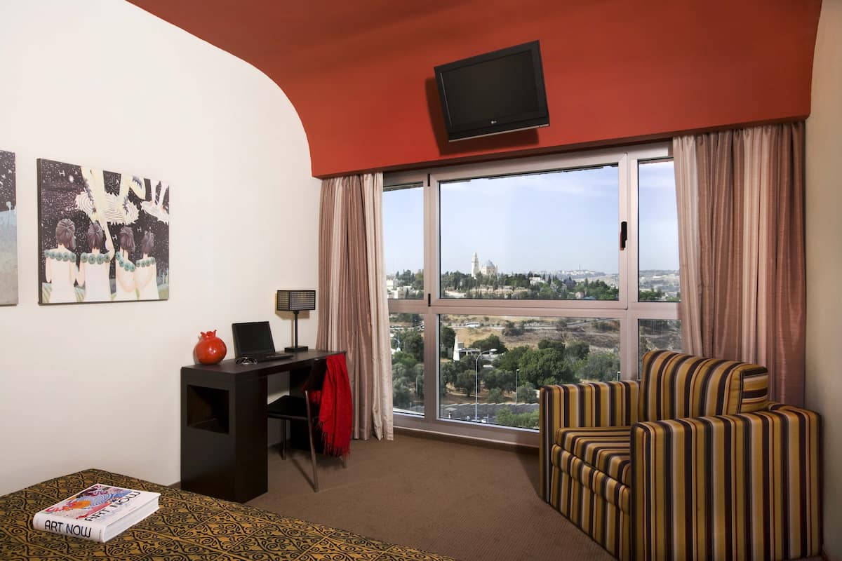 Une chambre d’hôtel avec une grande fenêtre, un bureau avec un ordinateur portable, un fauteuil à rayures et des œuvres d’art accrochées au mur.