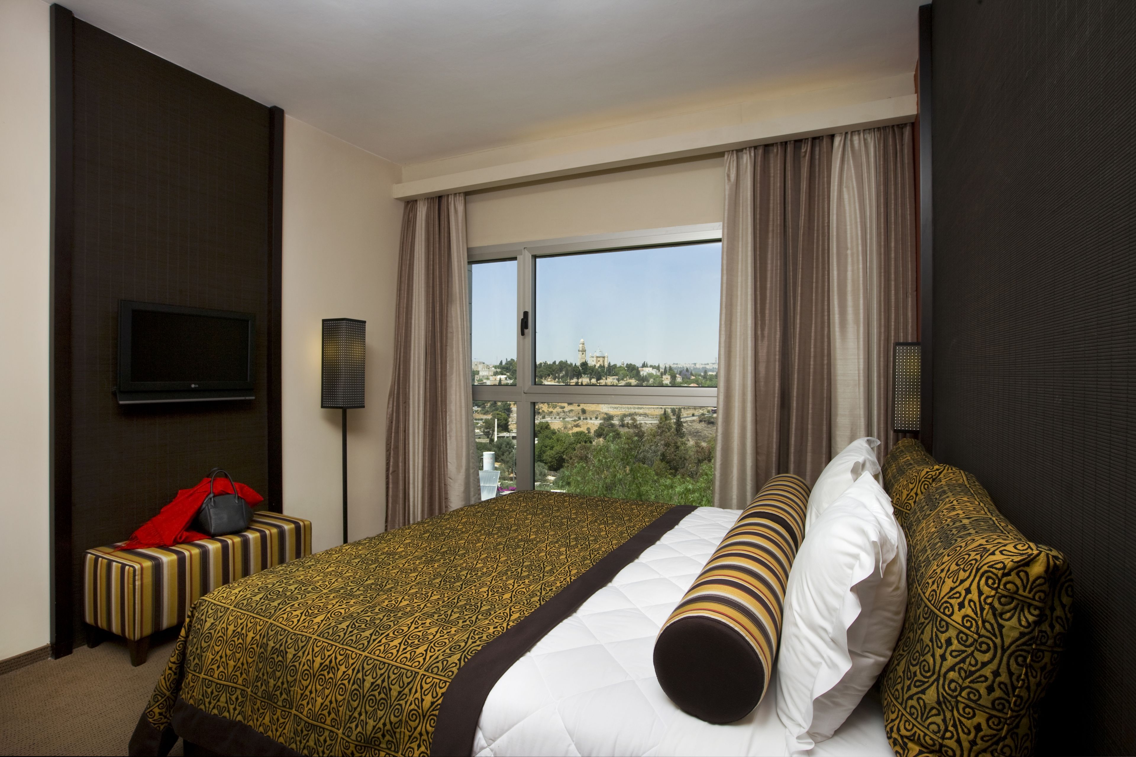 A hotel room with a large bed, a TV mounted on the wall, and a view of a cityscape through the window.