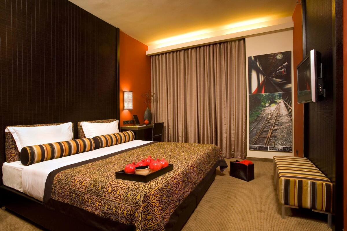 Une chambre d’hôtel avec un grand lit, une table de chevet avec une lampe, un petit bureau, une télévision et une fenêtre avec des rideaux.