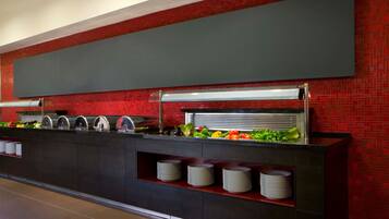 A buffet area with a red-tiled wall, a large flat-screen TV, and a variety of food items displayed on a counter.