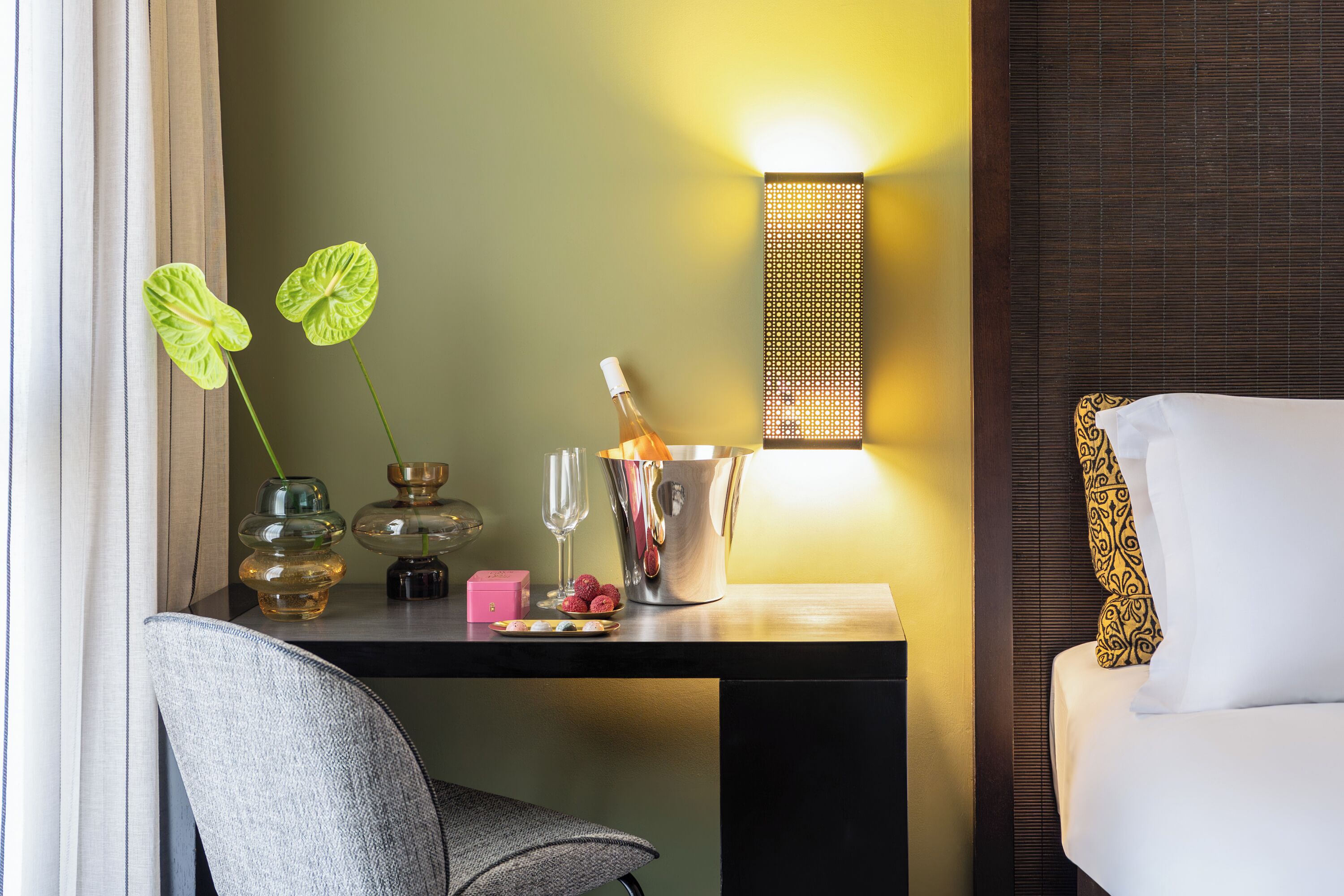 A hotel room with a wooden desk, a chair, a bed with patterned pillows, a wall-mounted light fixture, and a decorative plant.