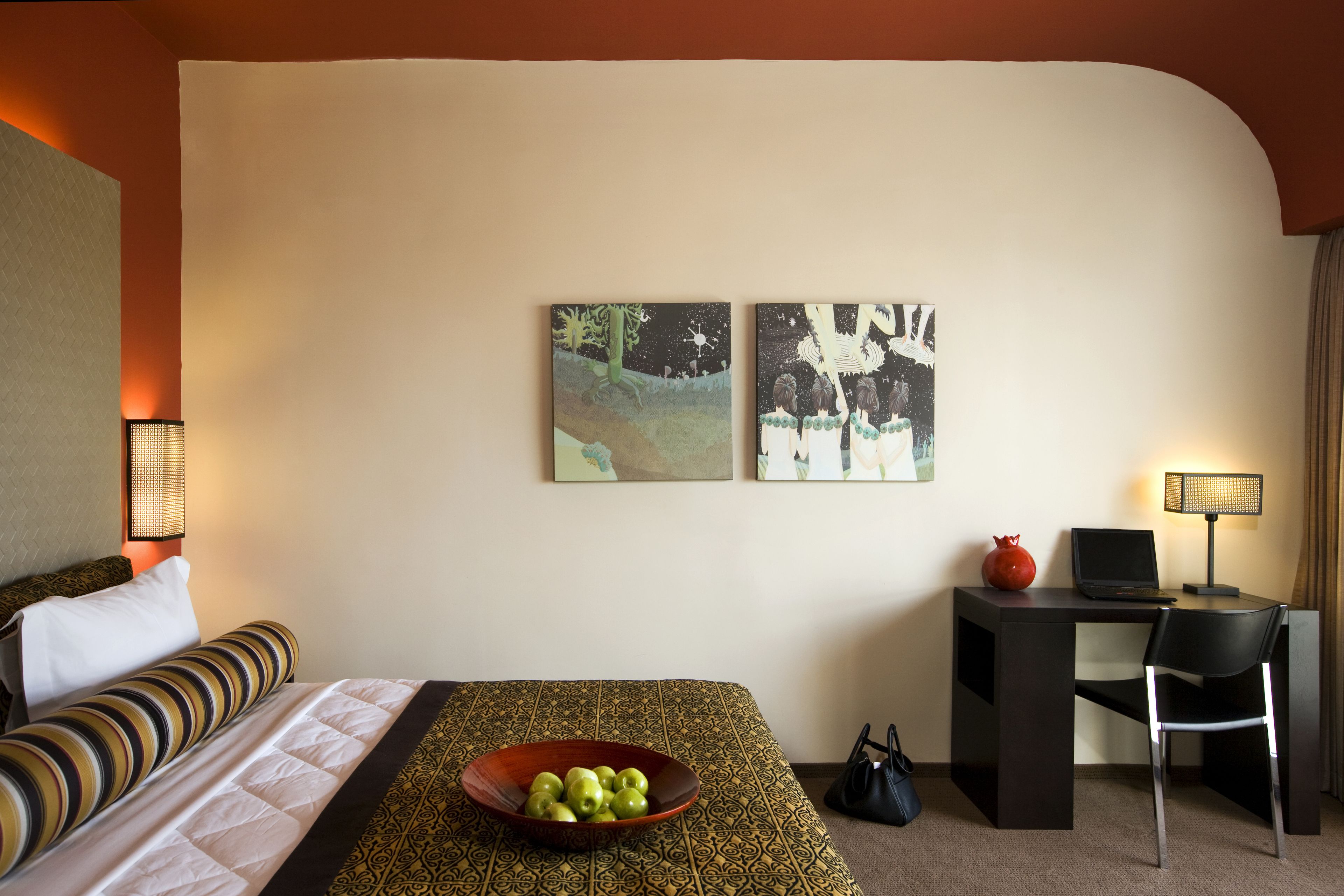 Quarto de hotel com cama, escrivaninha, laptop, uma tigela de fruta e duas pinturas na parede.