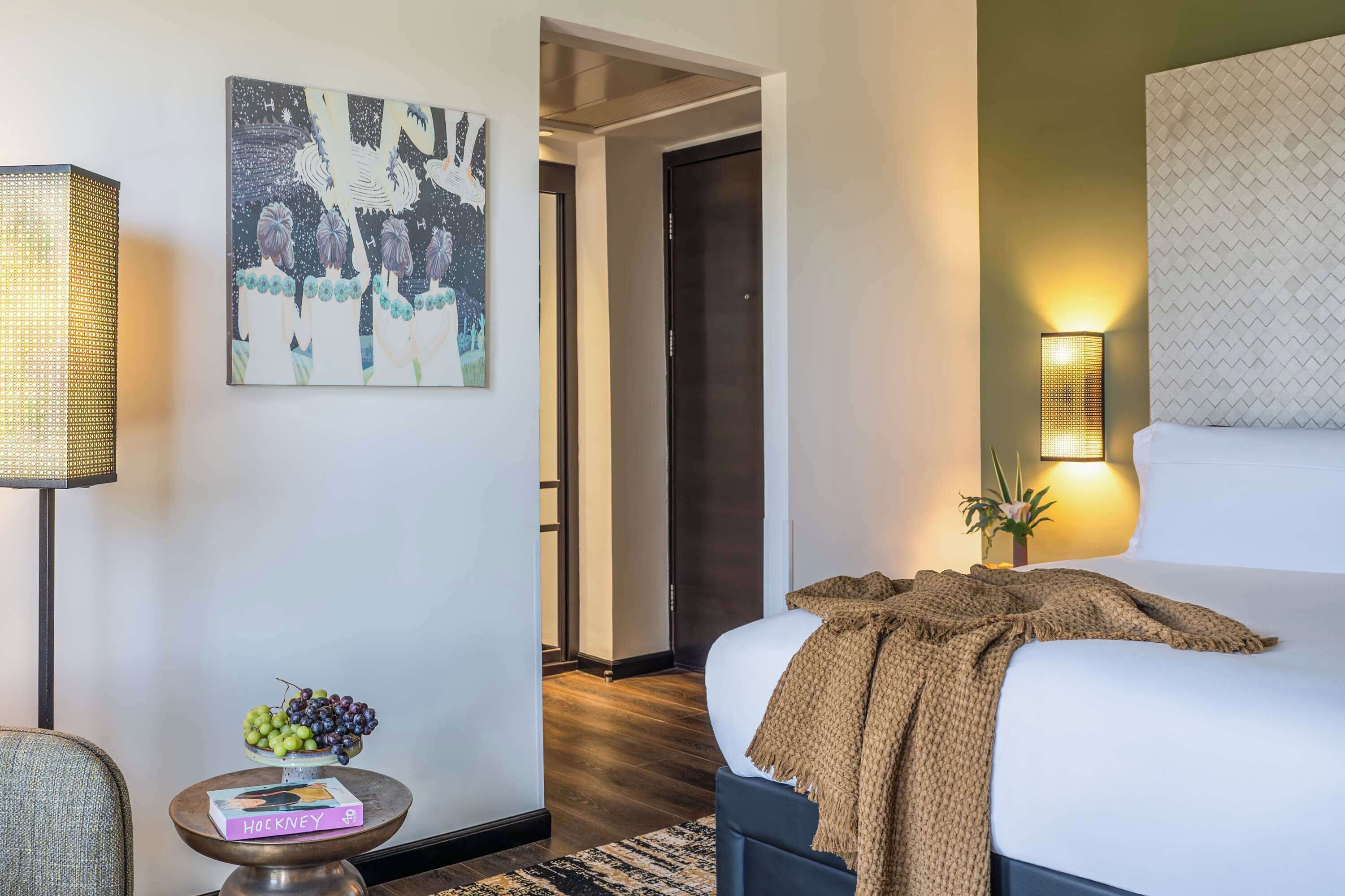 Quarto de hotel com uma cama, uma mesa de cabeceira com uma cesta de frutas, uma luminária, um quadro na parede e uma porta que dá para outro cômodo.