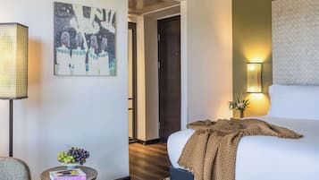 Quarto de hotel com uma cama, uma mesa de cabeceira com uma cesta de frutas, uma luminária, um quadro na parede e uma porta que dá para outro cômodo.