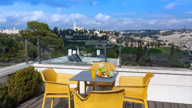 A terrace with yellow wicker chairs and a table, overlooking a cityscape with a prominent tower. - Dan Boutique Jerusalem (Jerusalem)