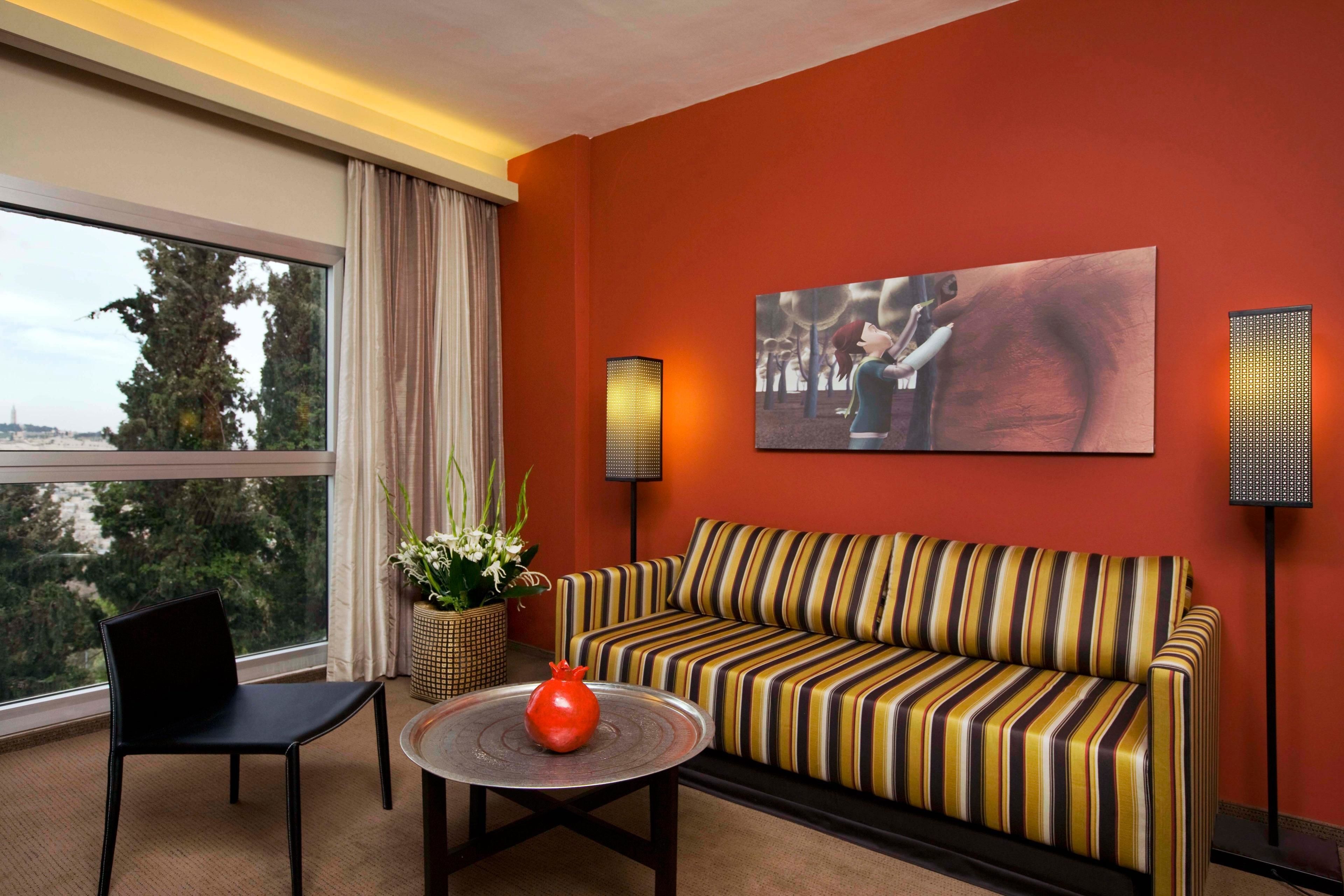 A modern living room with a striped sofa, a black chair, a round coffee table with a red vase, and a large window with a view of trees and a distant building.