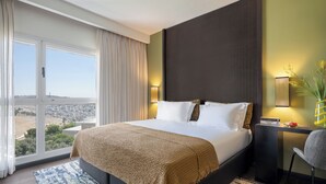 A hotel room with a large bed, a desk, a chair, and a view of the city through large windows. - Dan Boutique Jerusalem (Jerusalem)