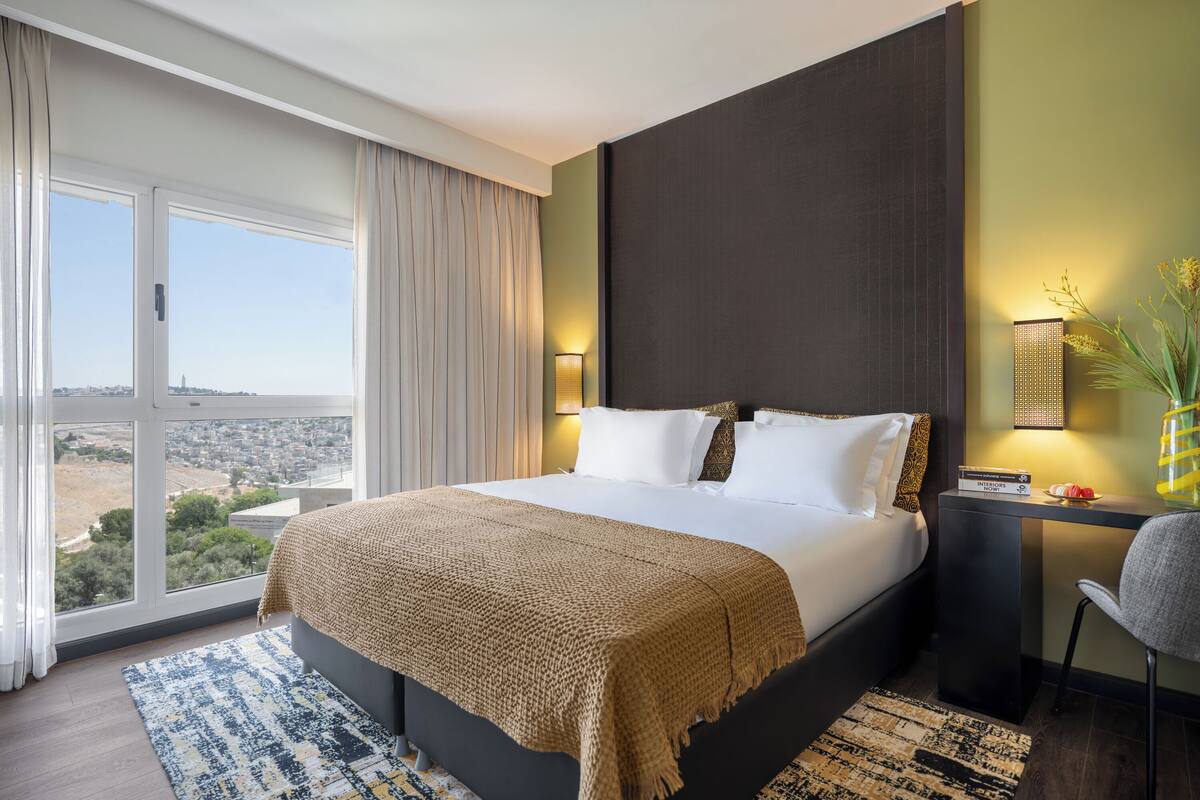 Une chambre d’hôtel avec un grand lit, un bureau, une chaise et une vue sur la ville grâce à de grandes fenêtres.