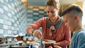 Iemand serveert eten aan een ander persoon in een keuken met betegelde muren en diverse kookgerei.