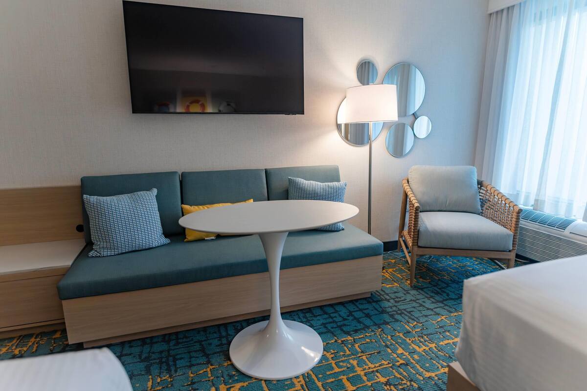 A hotel room with a sofa, a round table, a chair, a TV, and a lamp.