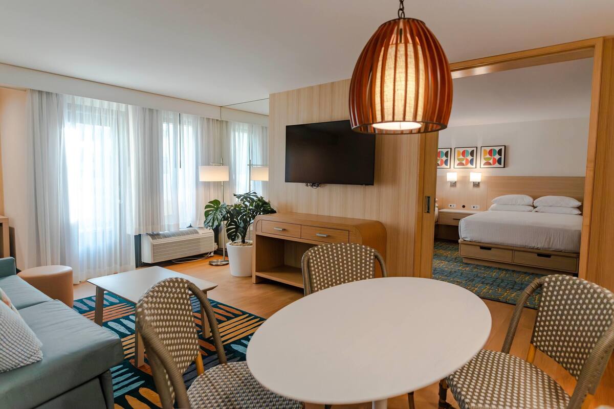 A hotel room with a sofa, dining table, chairs, a TV, a bed, a desk, a lamp, and a plant.