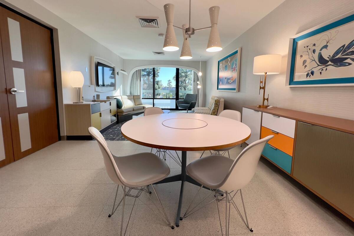A modern hotel room with a round dining table, two chairs, a sofa, a TV, and a large window.