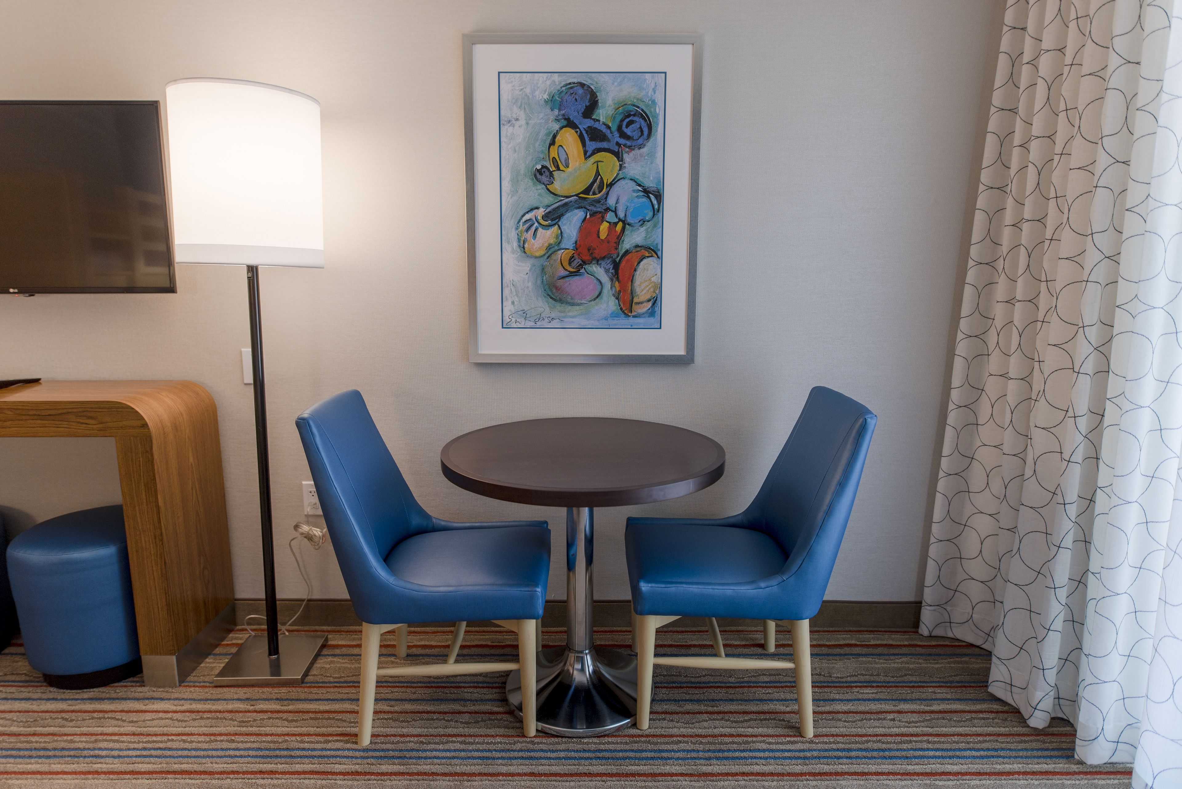 A hotel room with a round table, two blue chairs, a framed cartoon character artwork on the wall, a TV, and a patterned curtain.