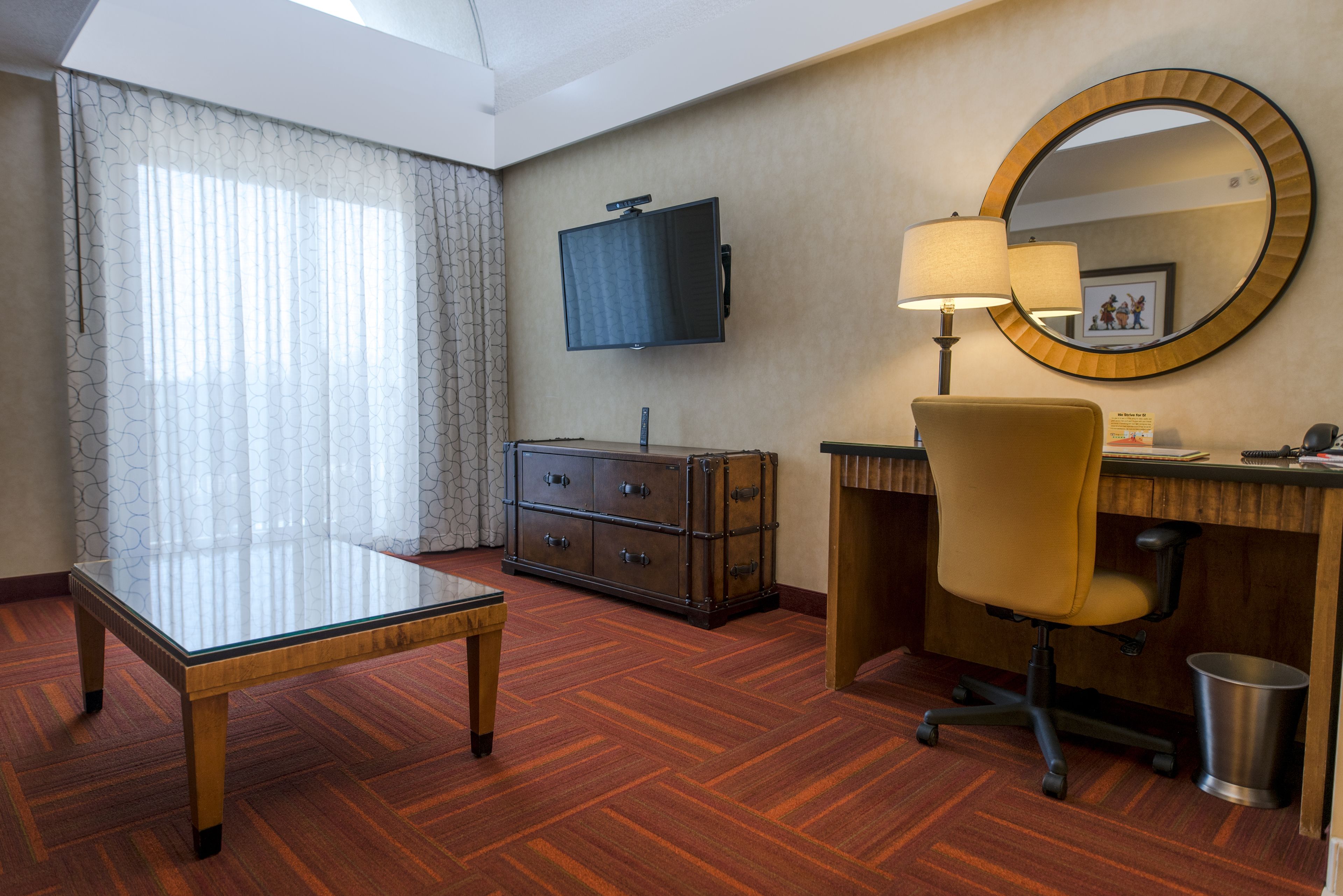 A hotel room with a wooden floor, a glass table, a desk with a chair, a television, a lamp, a mirror, and a trash can.
