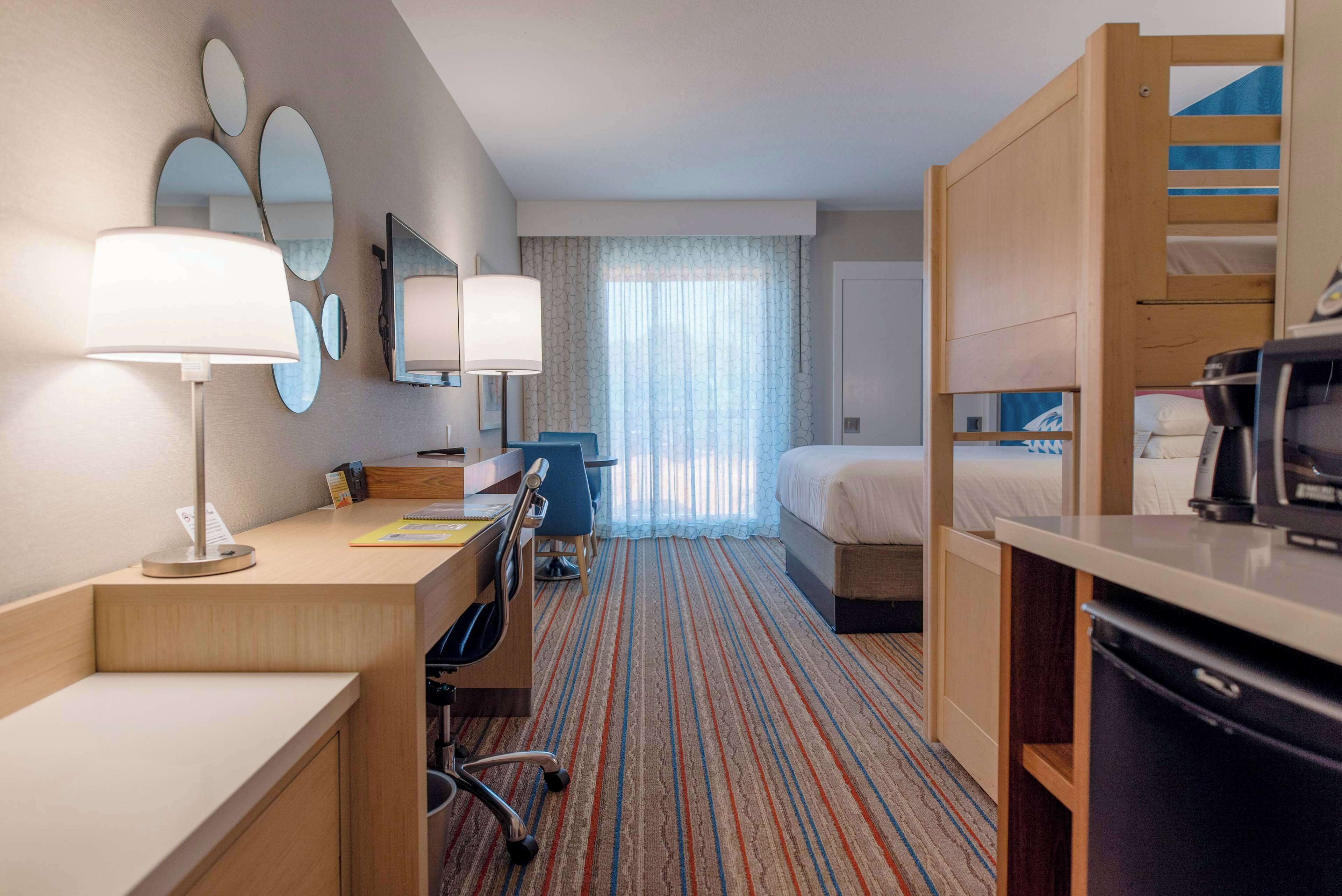 A hotel room with a desk, chair, bed, and a kitchenette.
