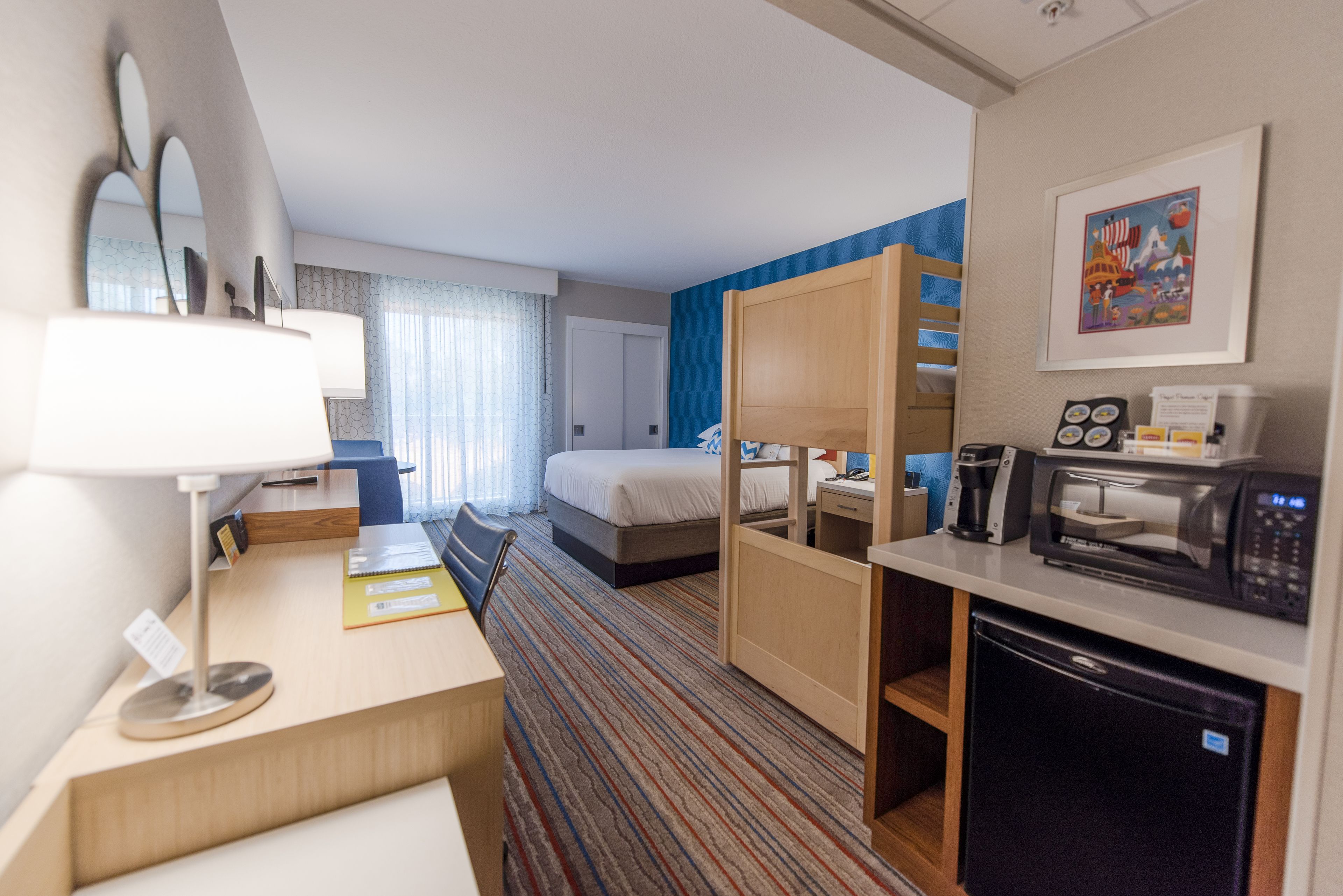 Hotellihuone, jossa on sänky, työpöytä lampun kanssa, mikroaaltouuni, jääkaappi ja siniraidallinen matto.