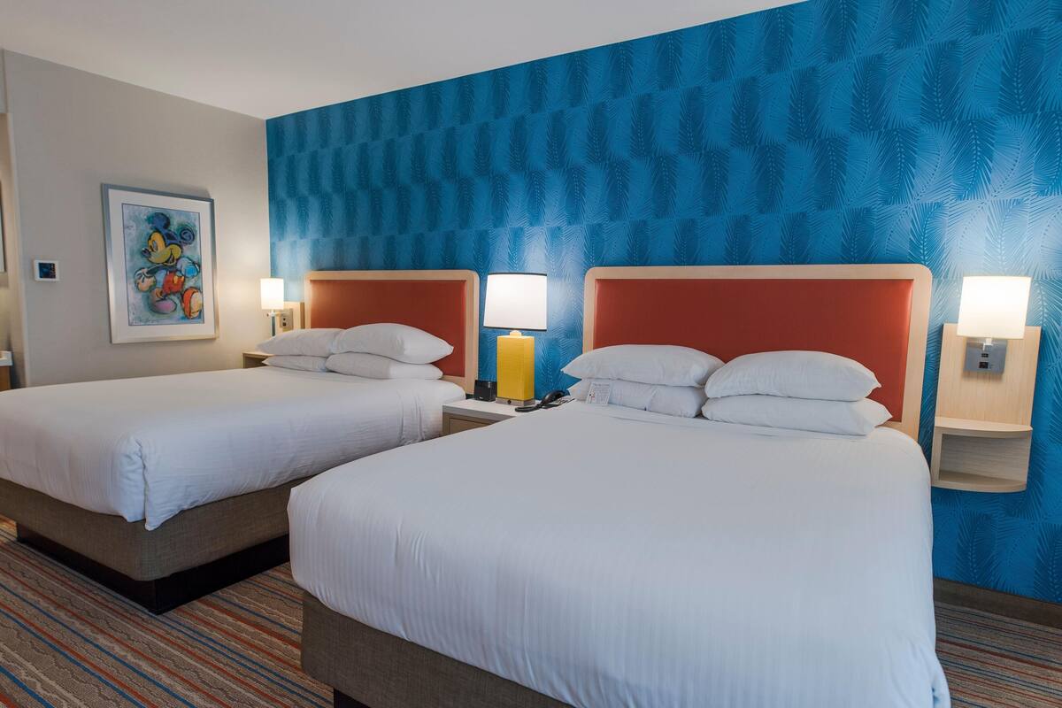 A hotel room with two beds, a blue patterned wall, and a Mickey Mouse painting.