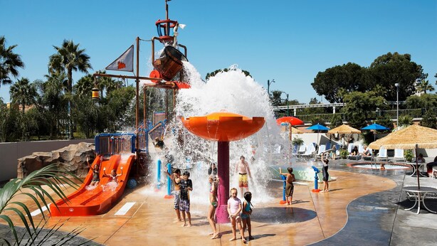 A water park with a water slide, a large water fountain, and a pool area with umbrellas. - Howard Johnson by Wyndham Anaheim Hotel and Water Playground (Anaheim)