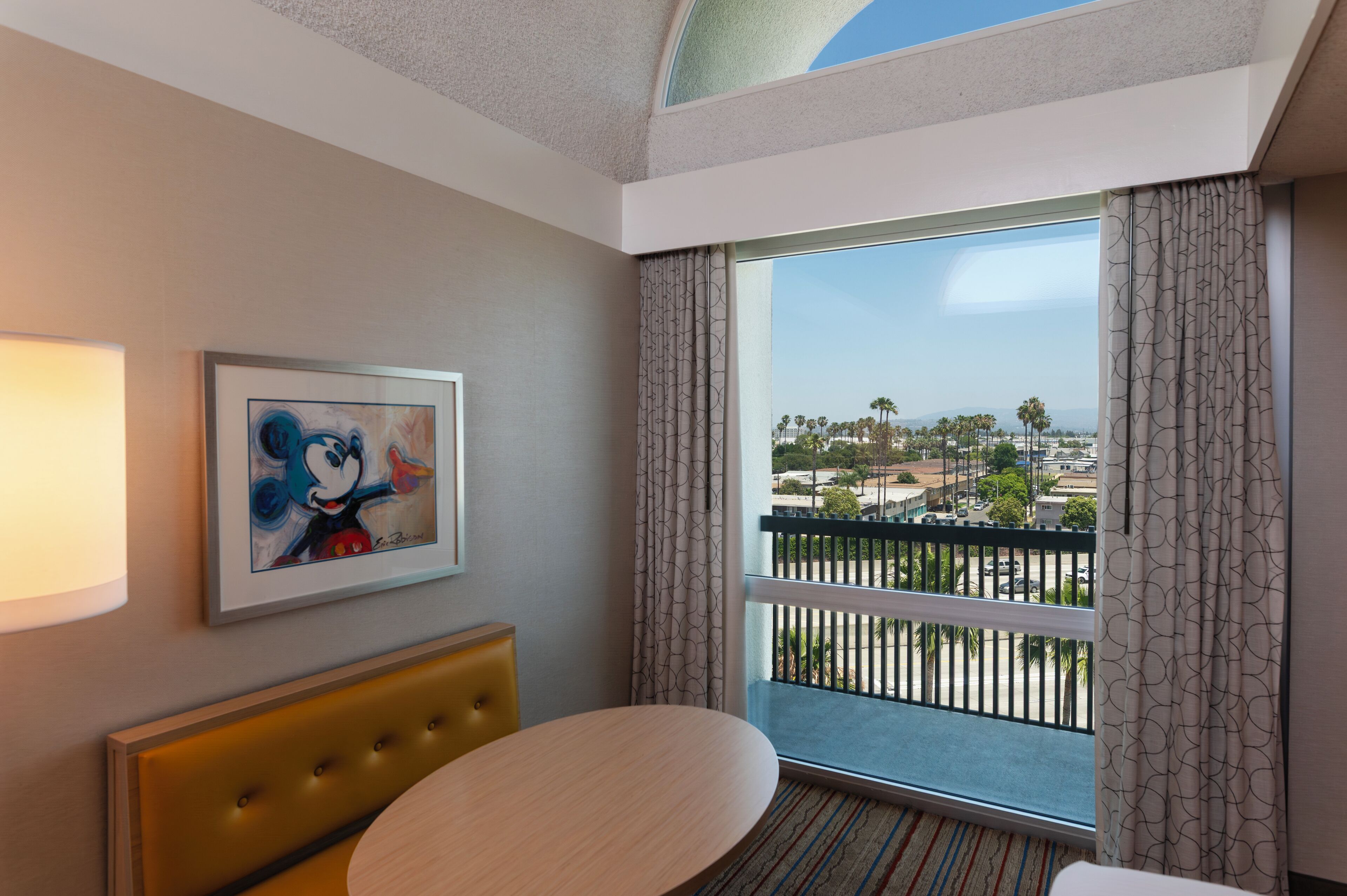 A hotel room with a large window, a framed artwork, a round table, and a tufted headboard.