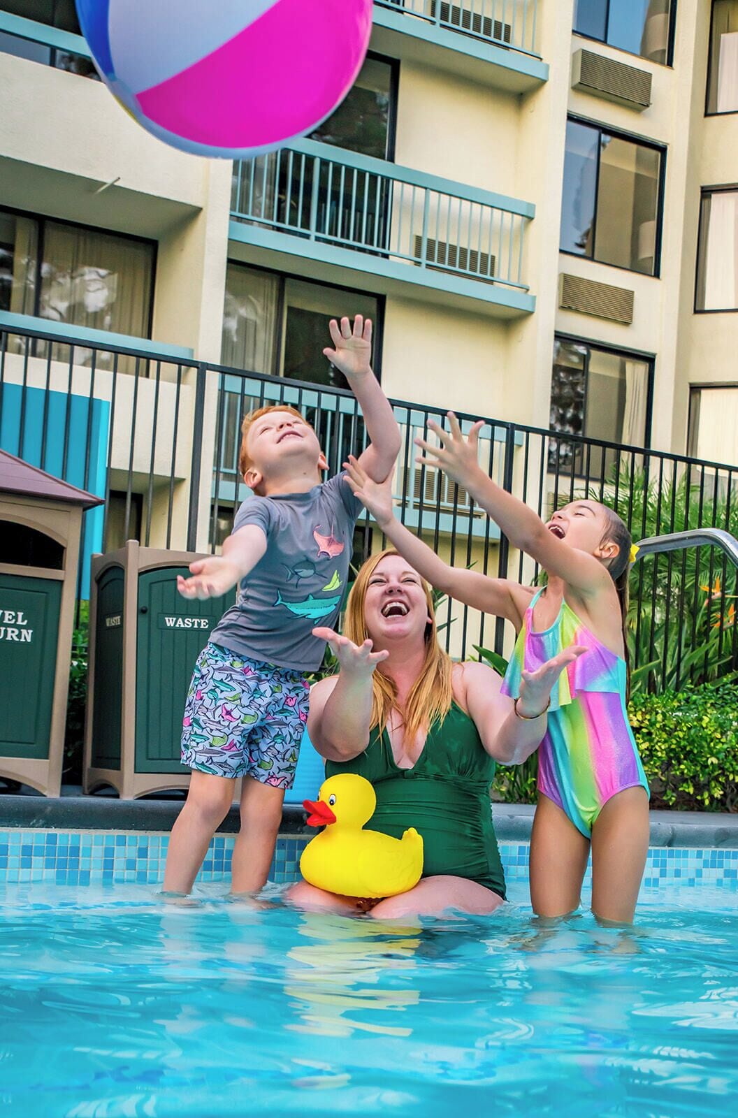 A swimming pool with a pink and white ball, a duck float, and people playing.
