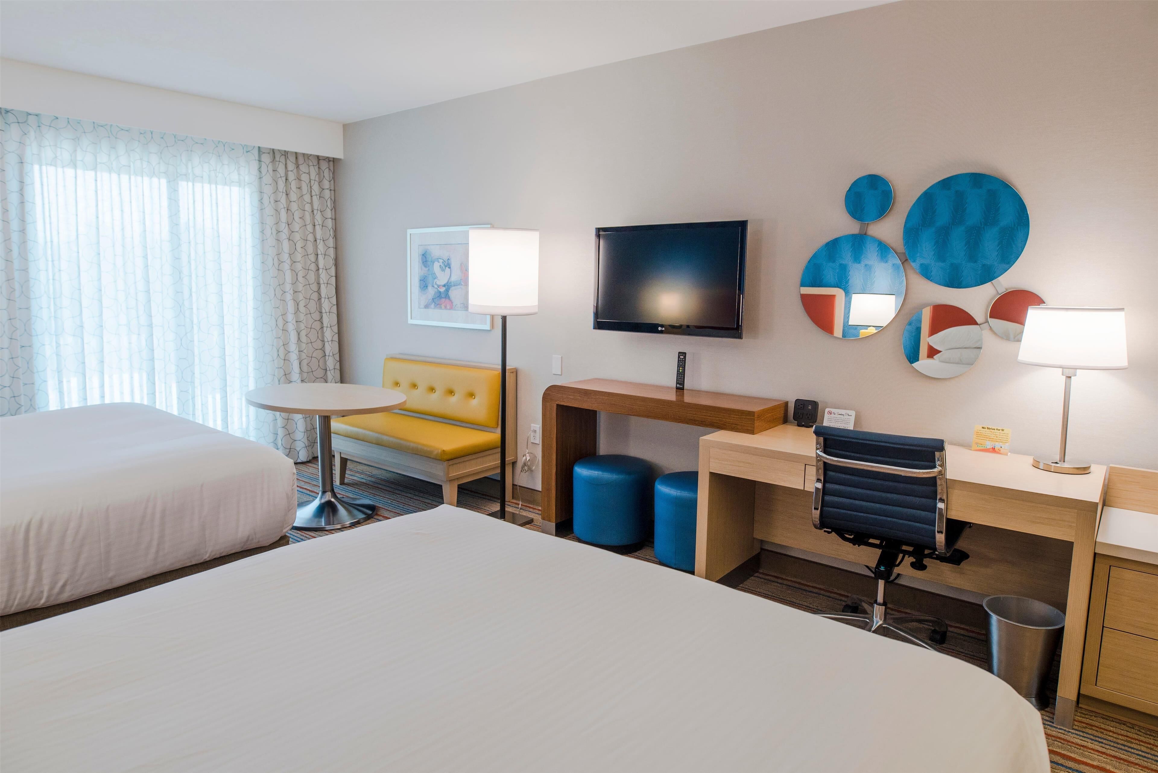 Une chambre d’hôtel avec deux lits, un bureau, une chaise, une télévision et un mur décoré d’œuvres d’art abstraites.