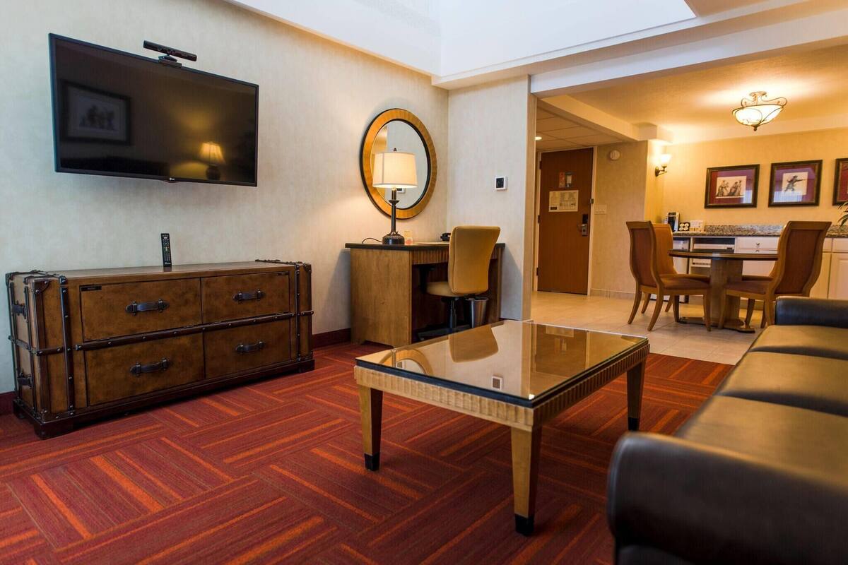 A hotel room with a flat-screen TV, a wooden cabinet, a glass coffee table, a sofa, and a dining area with chairs and a table.