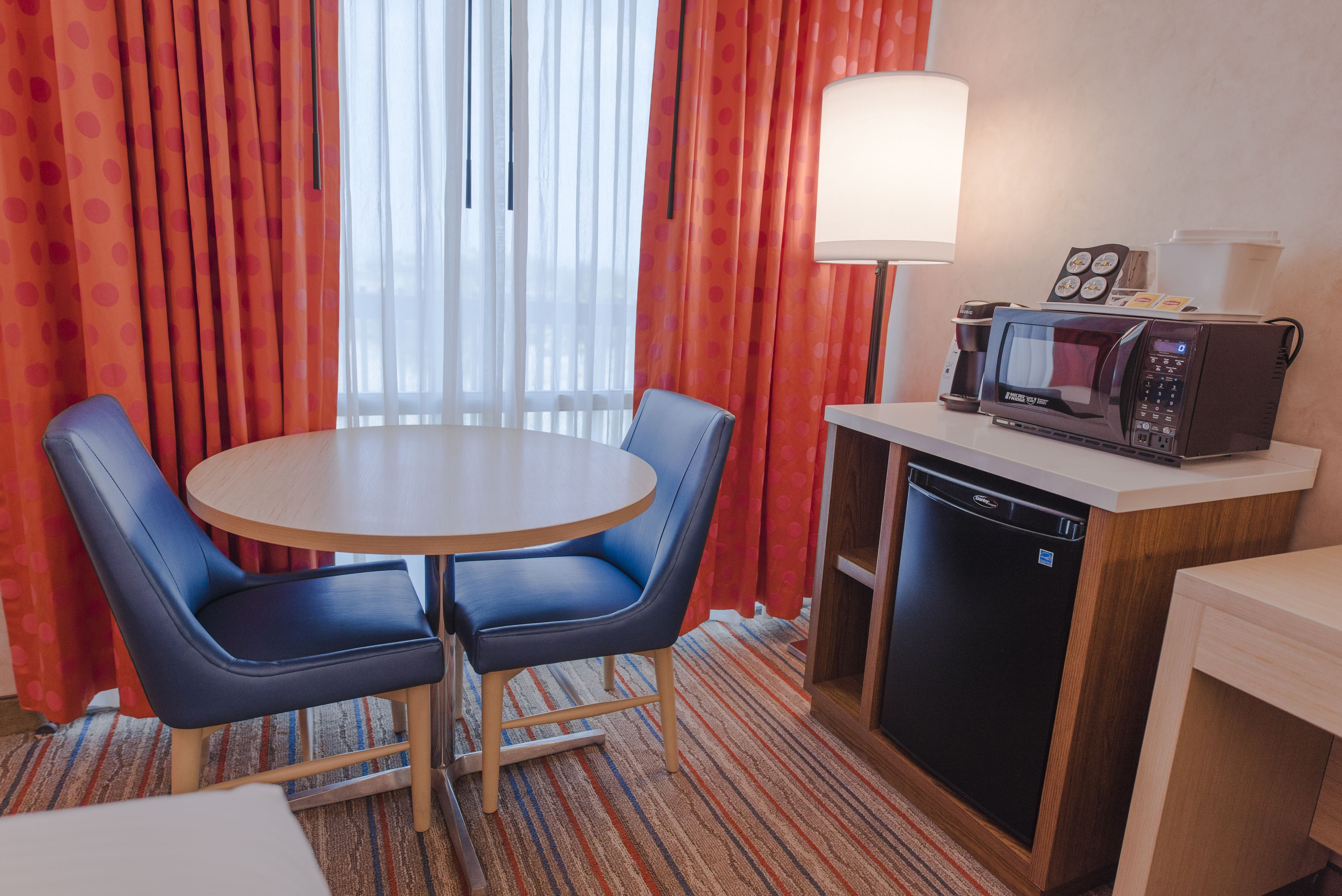 Ein Hotelzimmer mit einem runden Tisch, zwei blauen Stühlen, einer Lampe, einer Mikrowelle und einem Geschirrspüler.