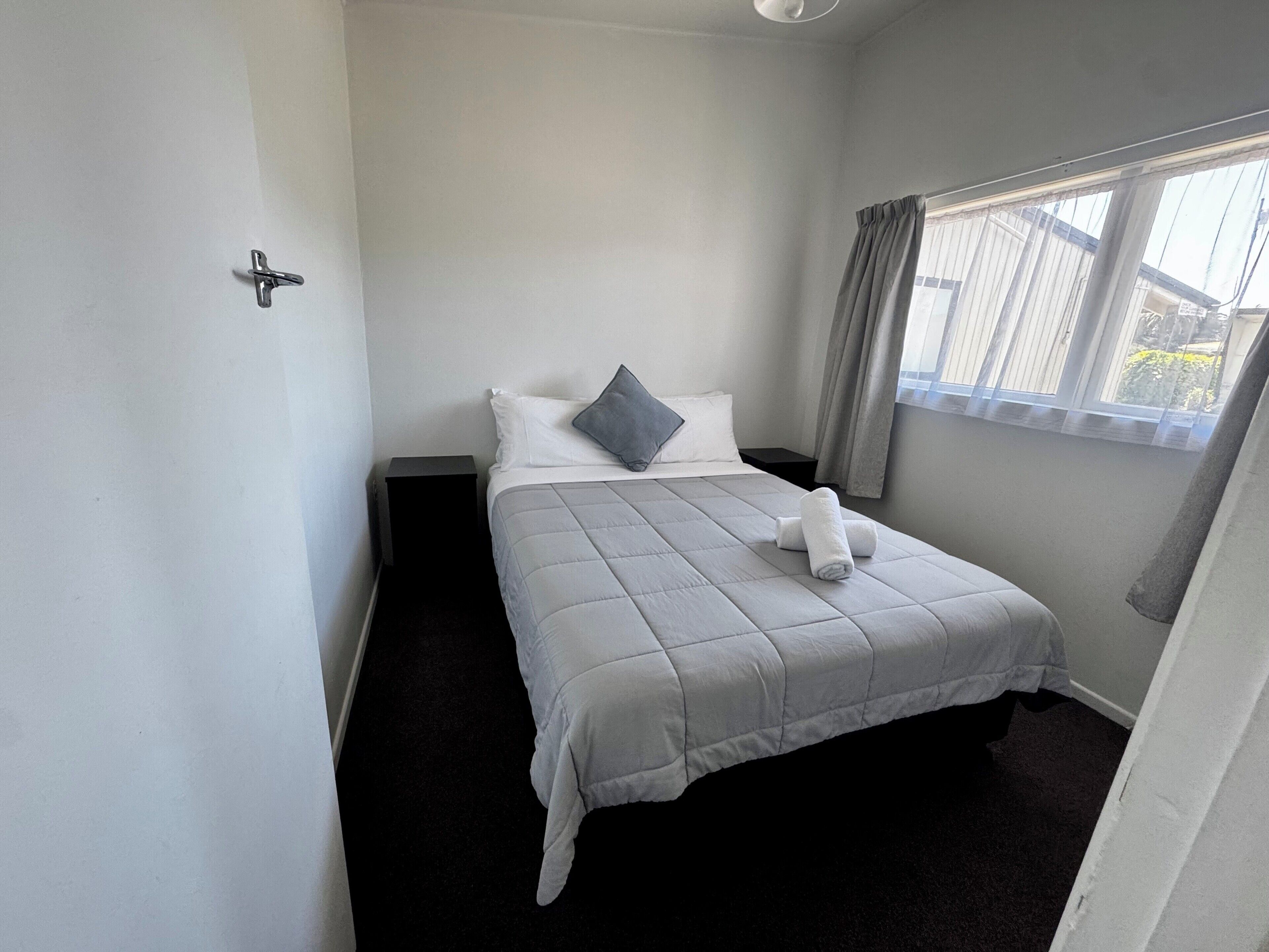Un lit bien fait, avec une couverture grise, un oreiller et une serviette enroulée. Il y a une fenêtre avec des rideaux, une petite table de chevet et un luminaire fixé au mur.