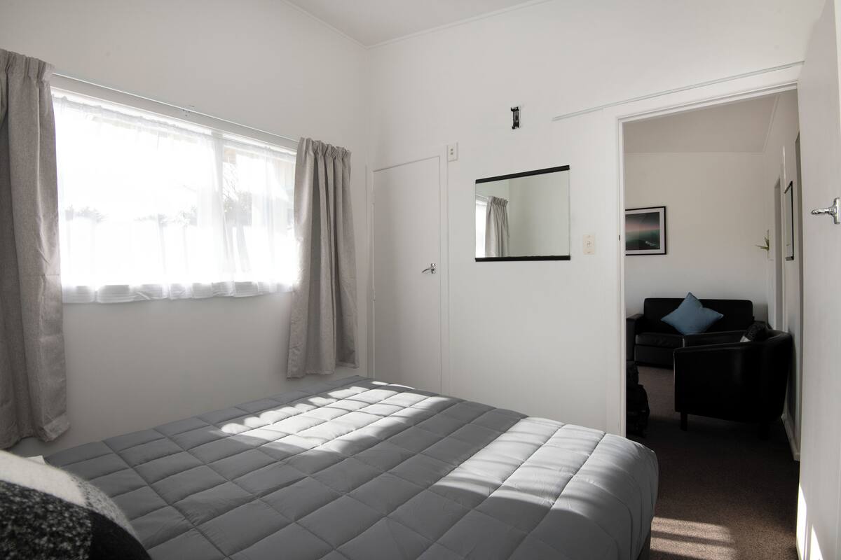 A bedroom with a bed, a window with curtains, a door, and a small seating area with a chair and a sofa.
