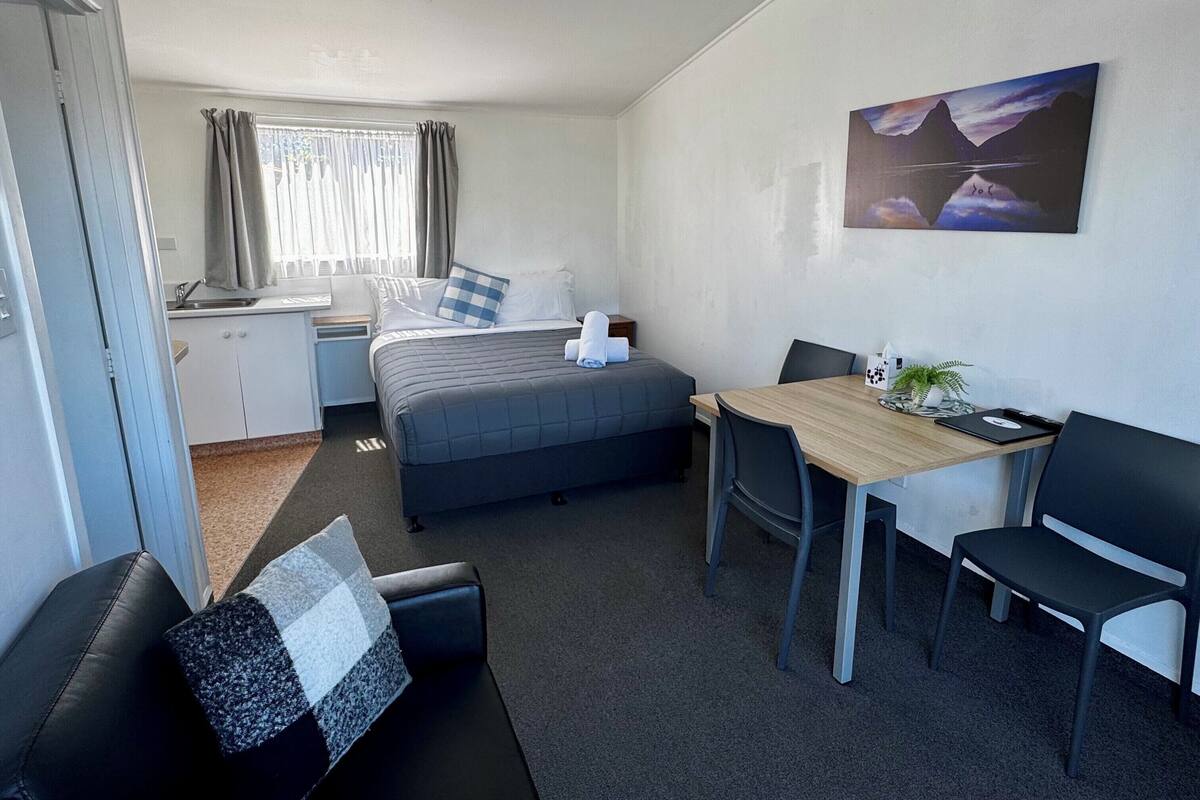A hotel room with a bed, a desk, a chair, a small table, and a painting on the wall.