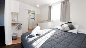 A hotel room with a bed, a bedside table, a window with curtains, a bathroom with a sink and mirror, and a hallway leading to another room. - Spanish Lady Motel (Napier)
