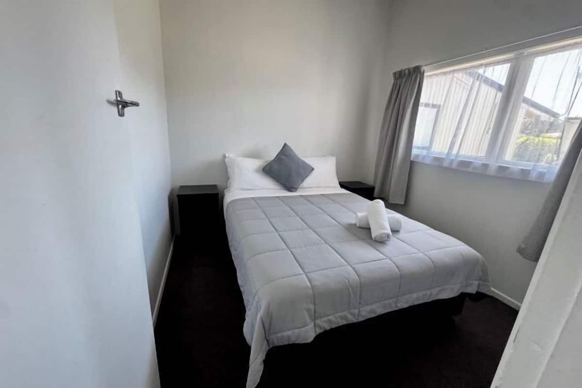 Un lit simple avec une couvre-lit blanche et un oreiller gris, une petite table de chevet et une fenêtre avec des rideaux.