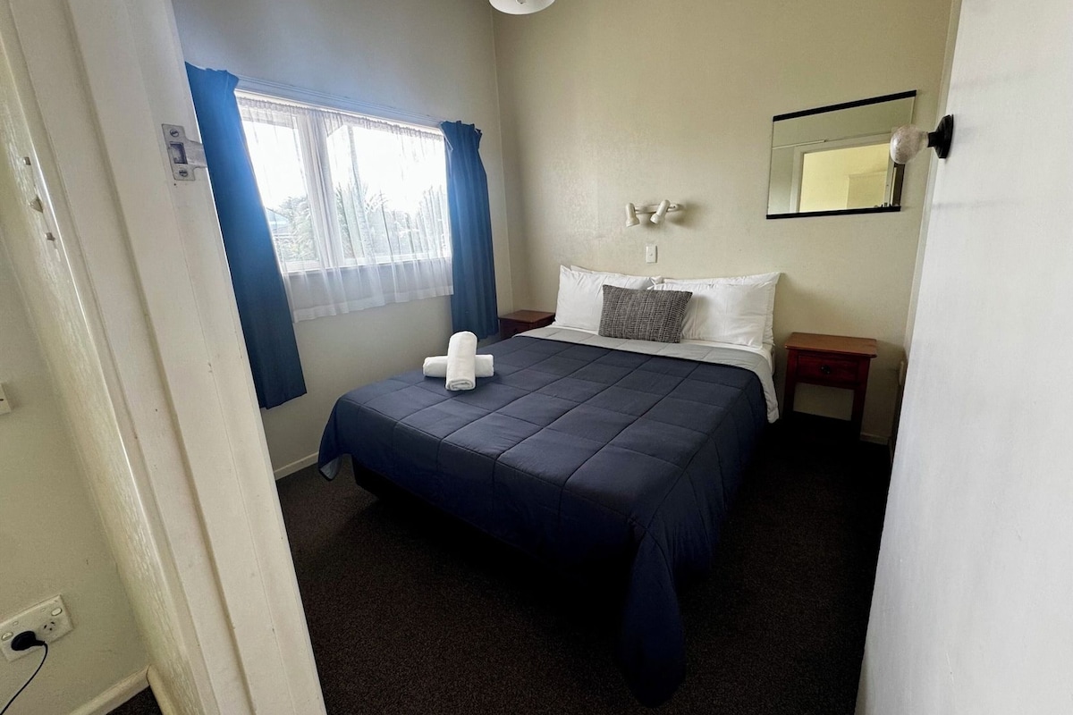 A bedroom with a bed, a bedside table, a nightstand, a window with curtains, and a wall-mounted light fixture.